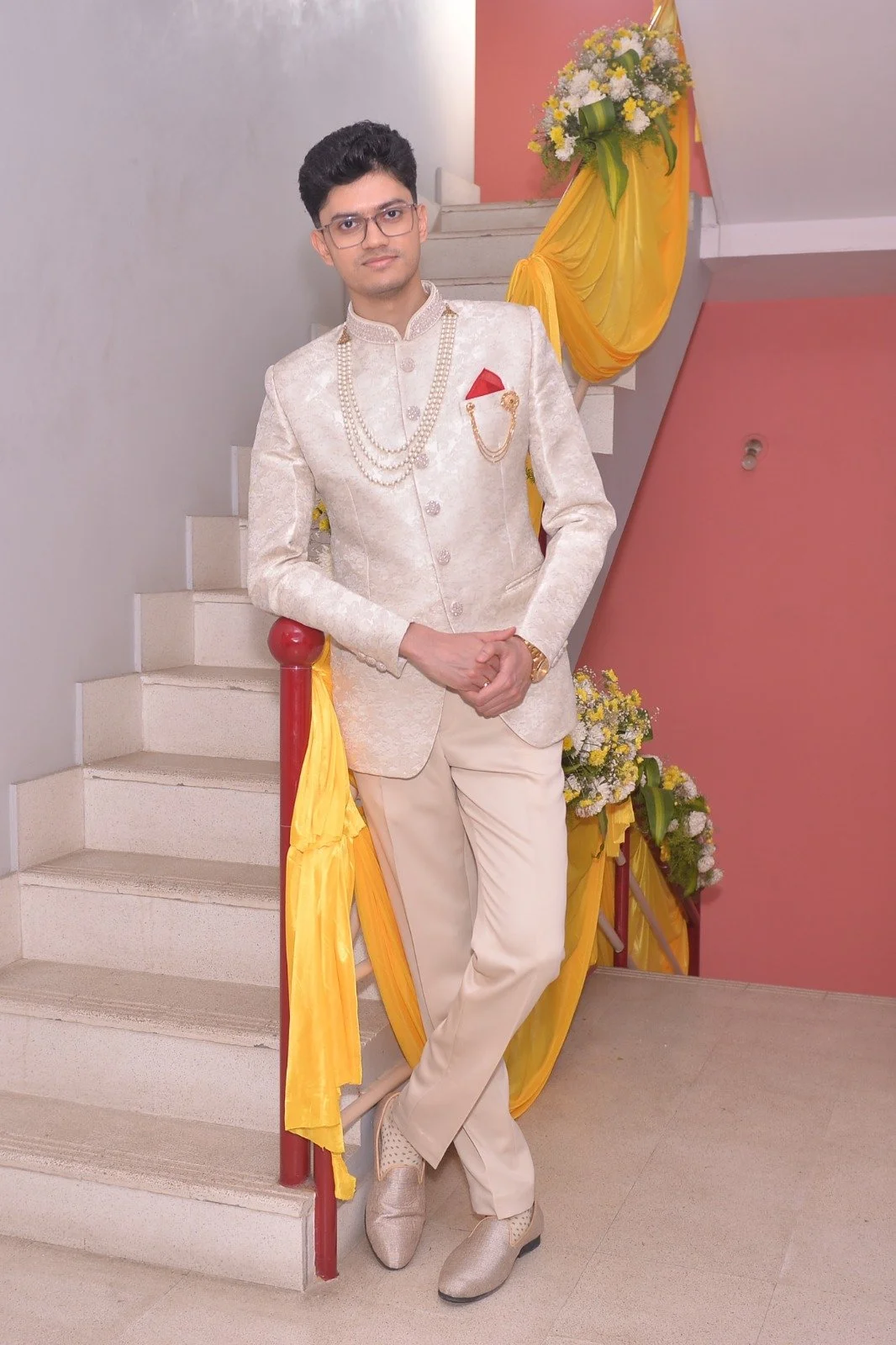 A young man in traditional cream-colored wedding attire standing on a staircase decorated with yellow and white flowers and yellow fabric.