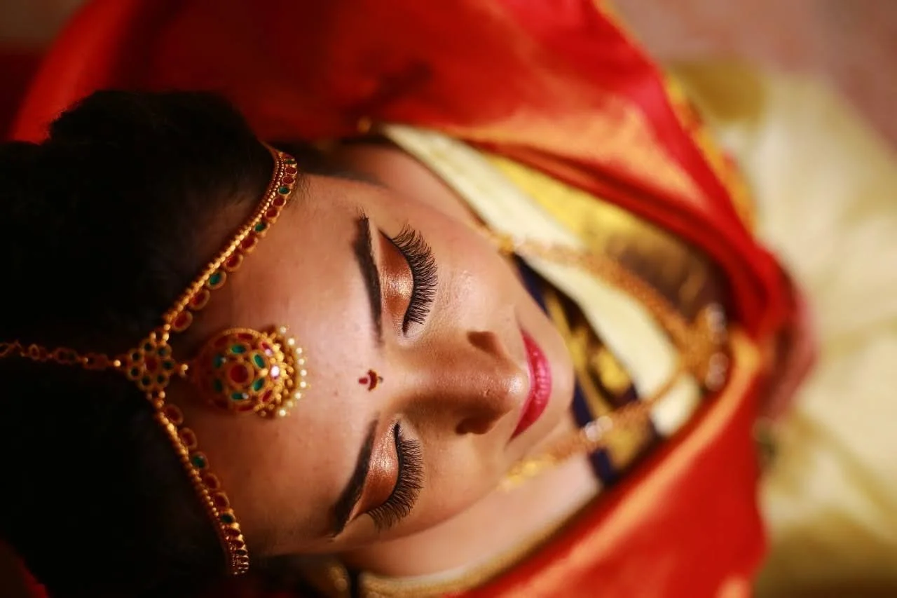 Close-up of a woman with makeup and traditional jewelry, lying down with her eyes closed, surrounded by red and yellow fabric.