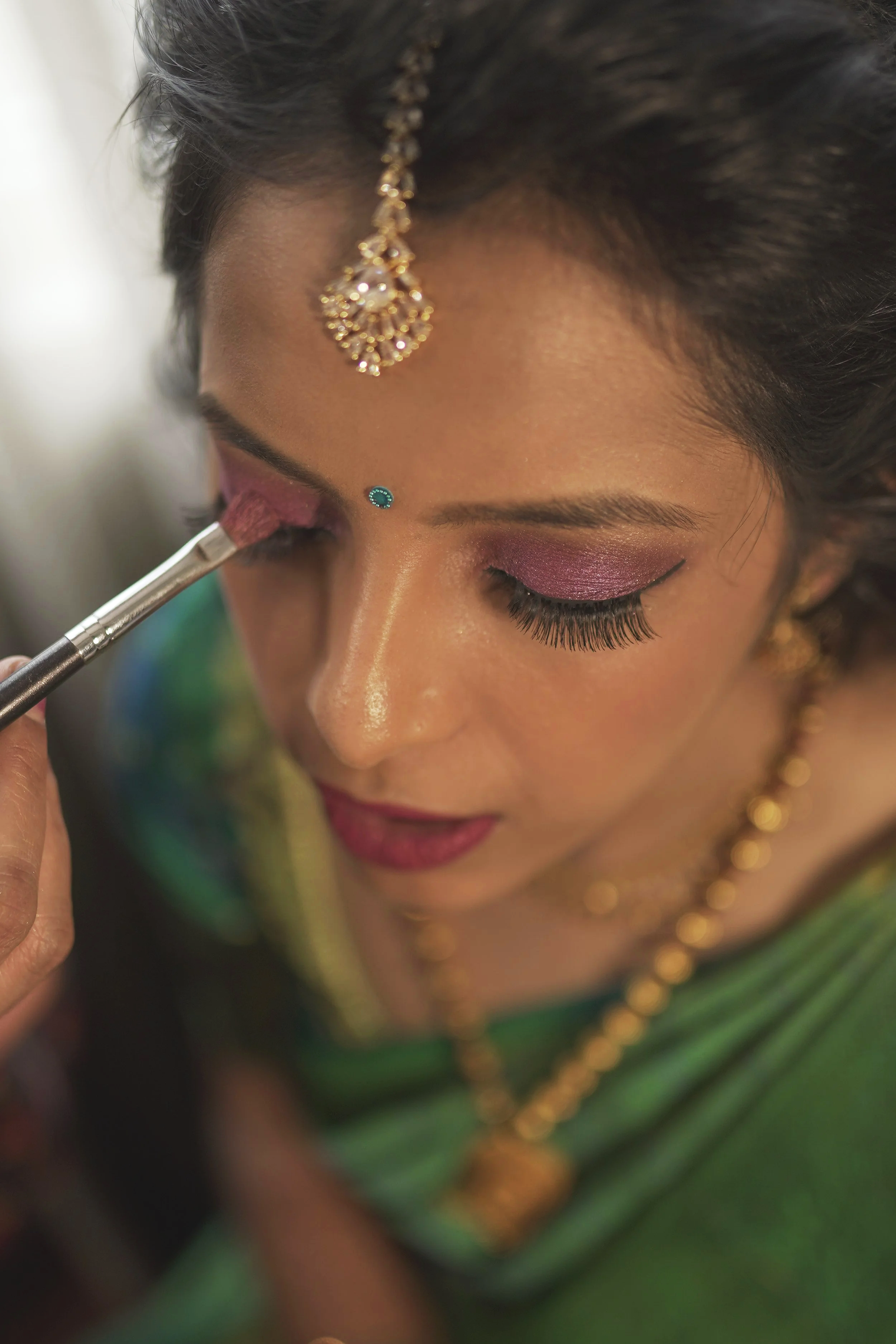 A woman with traditional Indian jewelry getting her eye makeup painted pink.