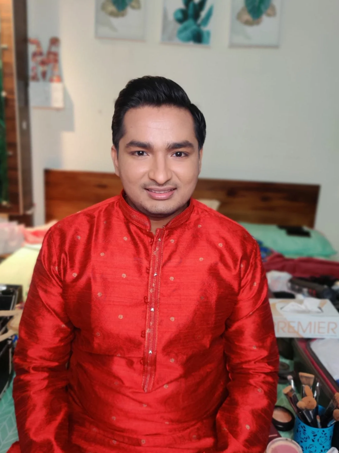 A man with dark hair, dressed in a red traditional Asian outfit, sitting indoors with a smile.