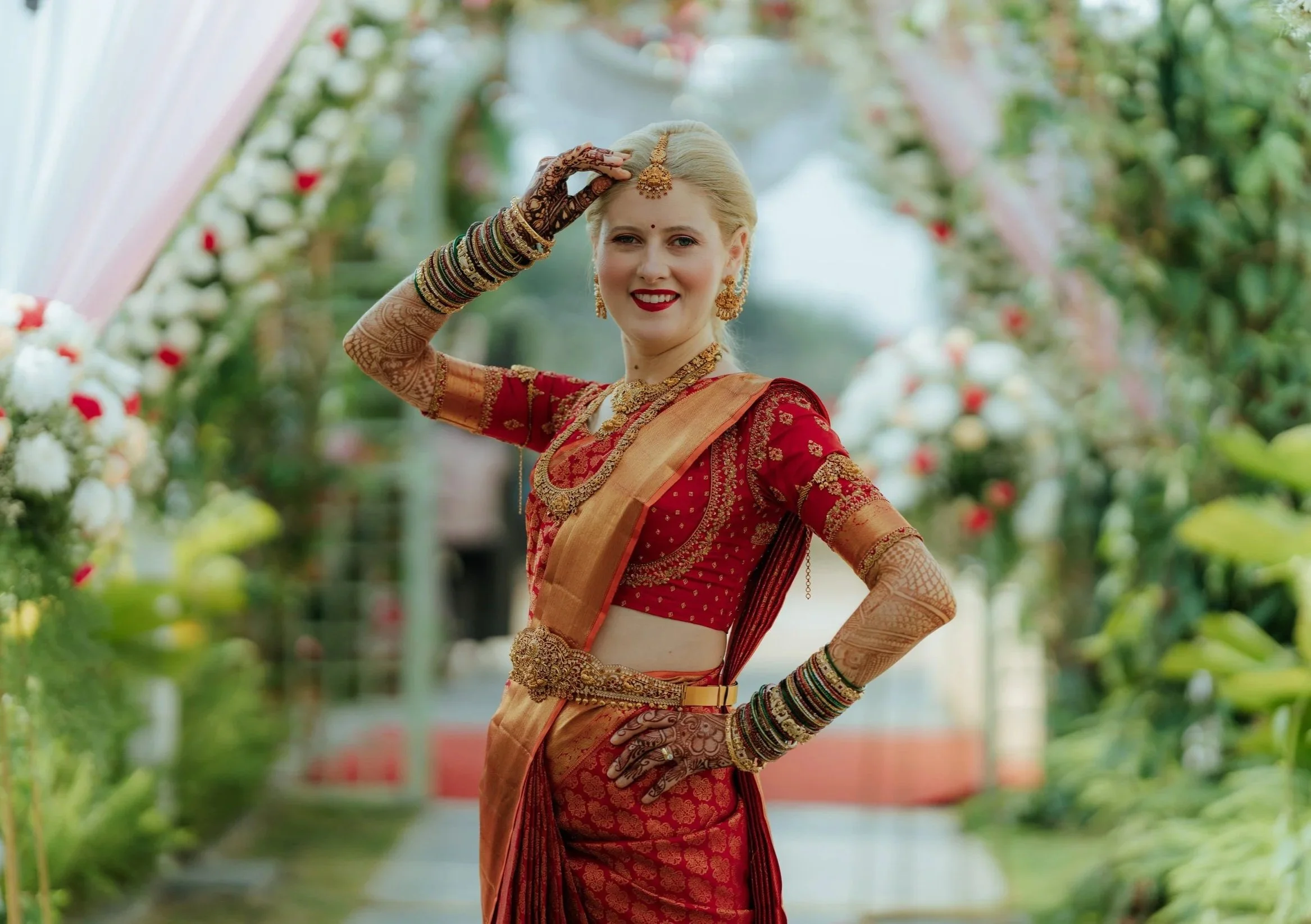 A woman dressed in traditional Indian bridal attire, wearing a red and gold sari, adorned with jewelry, bangles, and mehndi on her hands, standing outdoors with floral decorations in the background.