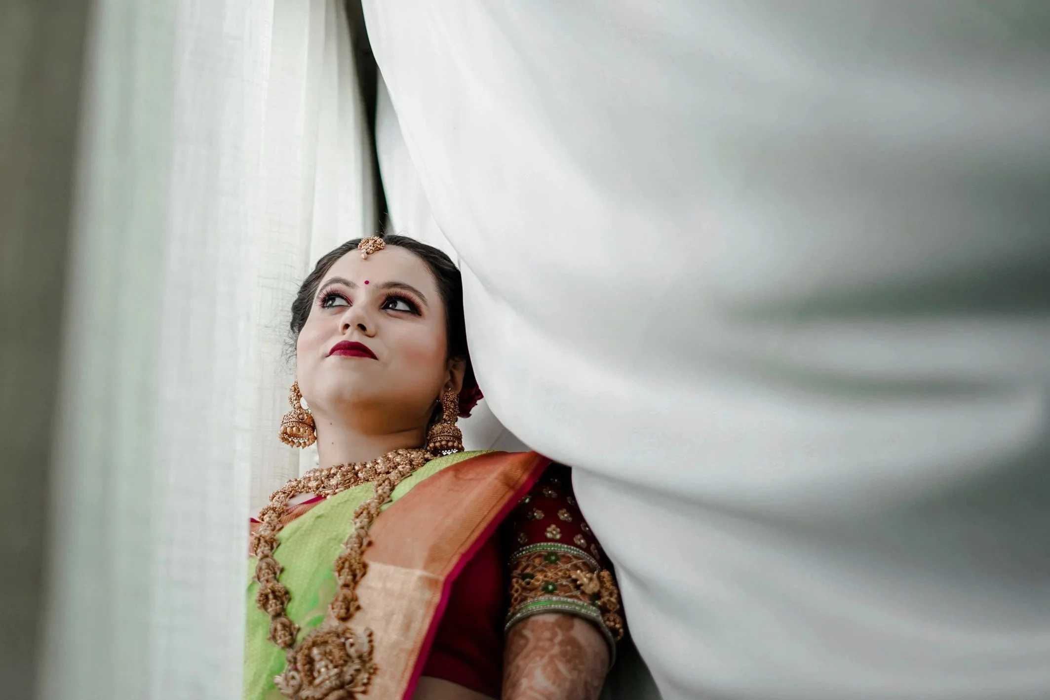 Young woman dressed in traditional Indian attire, wearing jewelry, looking upward with a contemplative expression, surrounded by white fabric or curtains.
