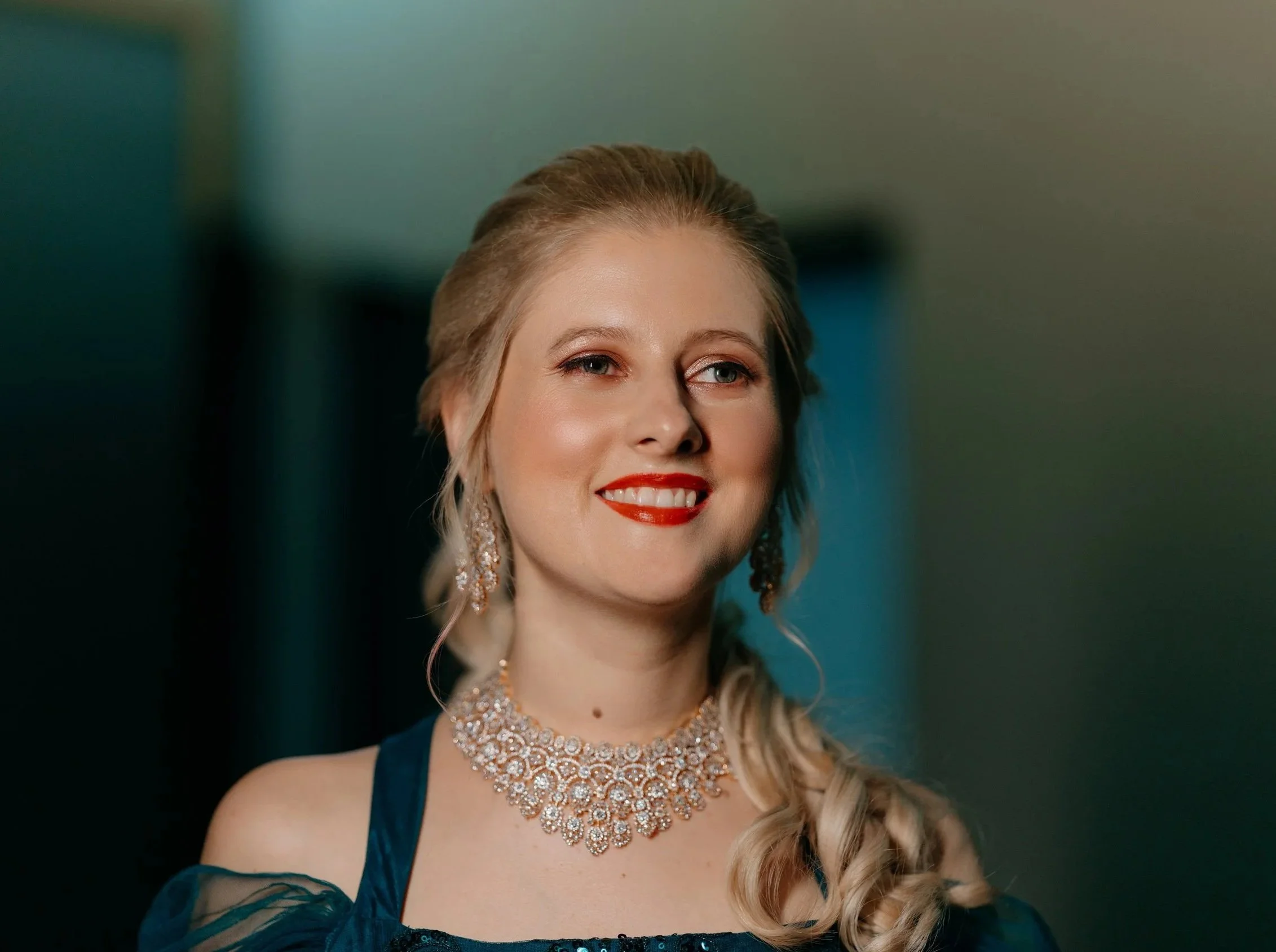 A woman with blonde hair styled in loose curls, wearing a dark blue dress with off-the-shoulder sleeves, and accessorized with large, sparkling earrings and a matching necklace, smiling with red lipstick.