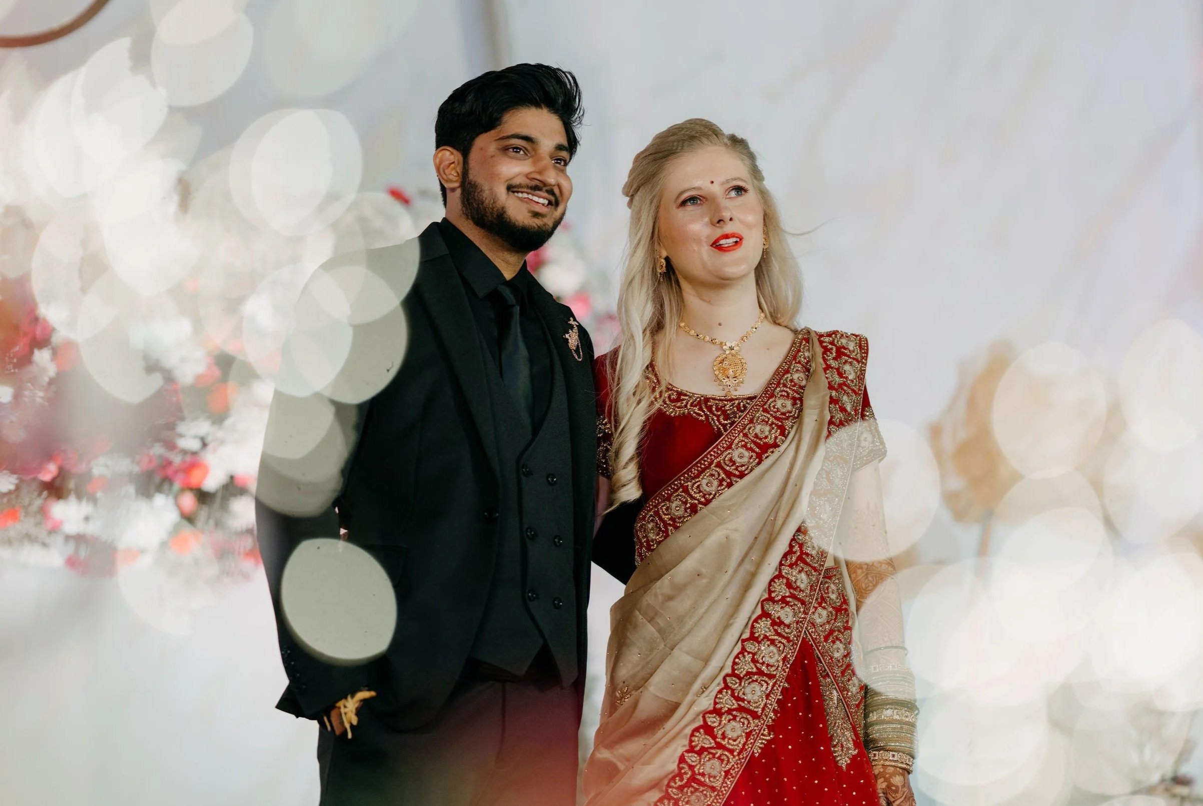 A couple dressed in traditional Indian attire stands close together, smiling at an event decorated with warm bokeh lights and floral arrangements.