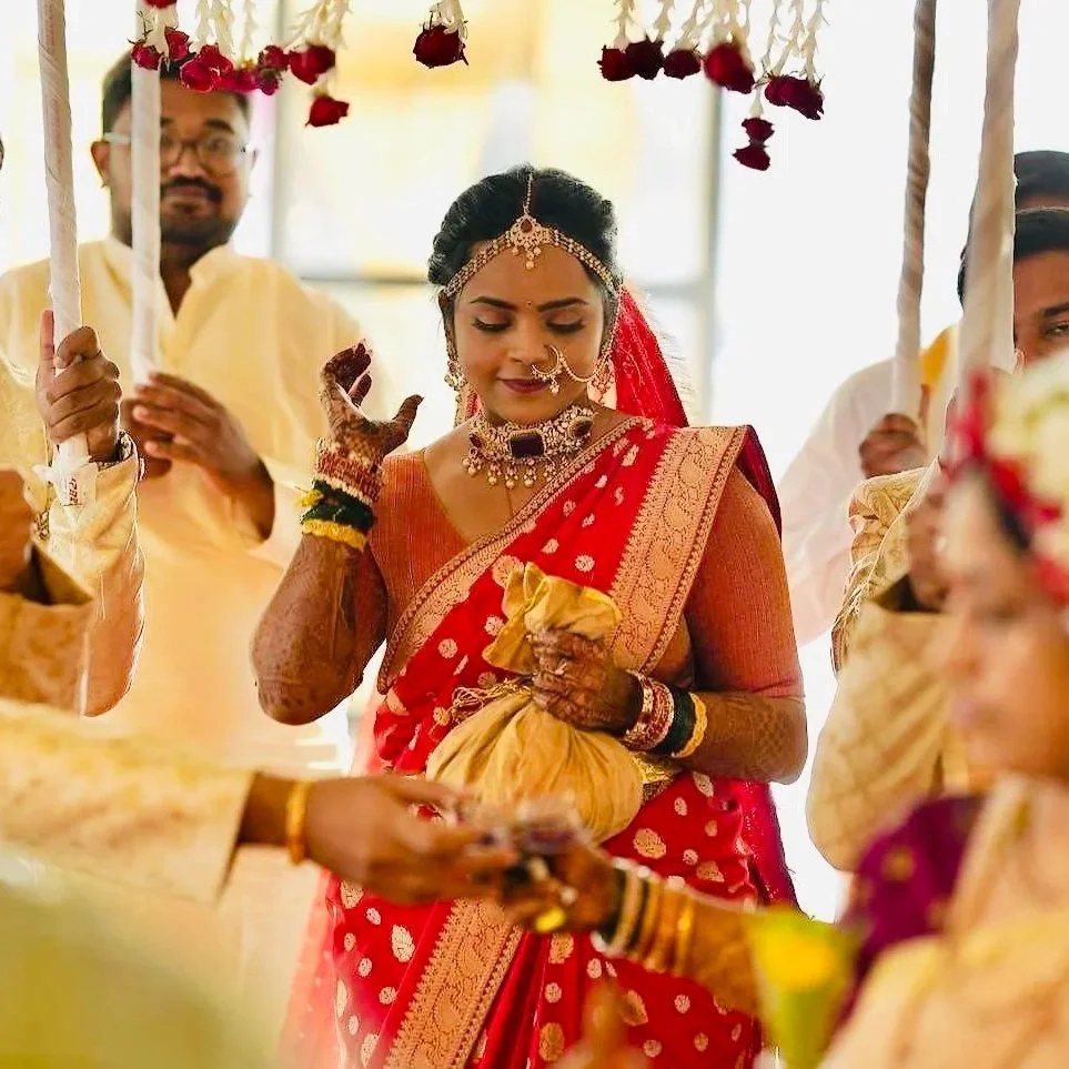 An Indian woman dressed in traditional red and gold bridal attire, wearing jewelry, including a nose ring, necklace, and headpiece, during a Hindu wedding ceremony among family members.