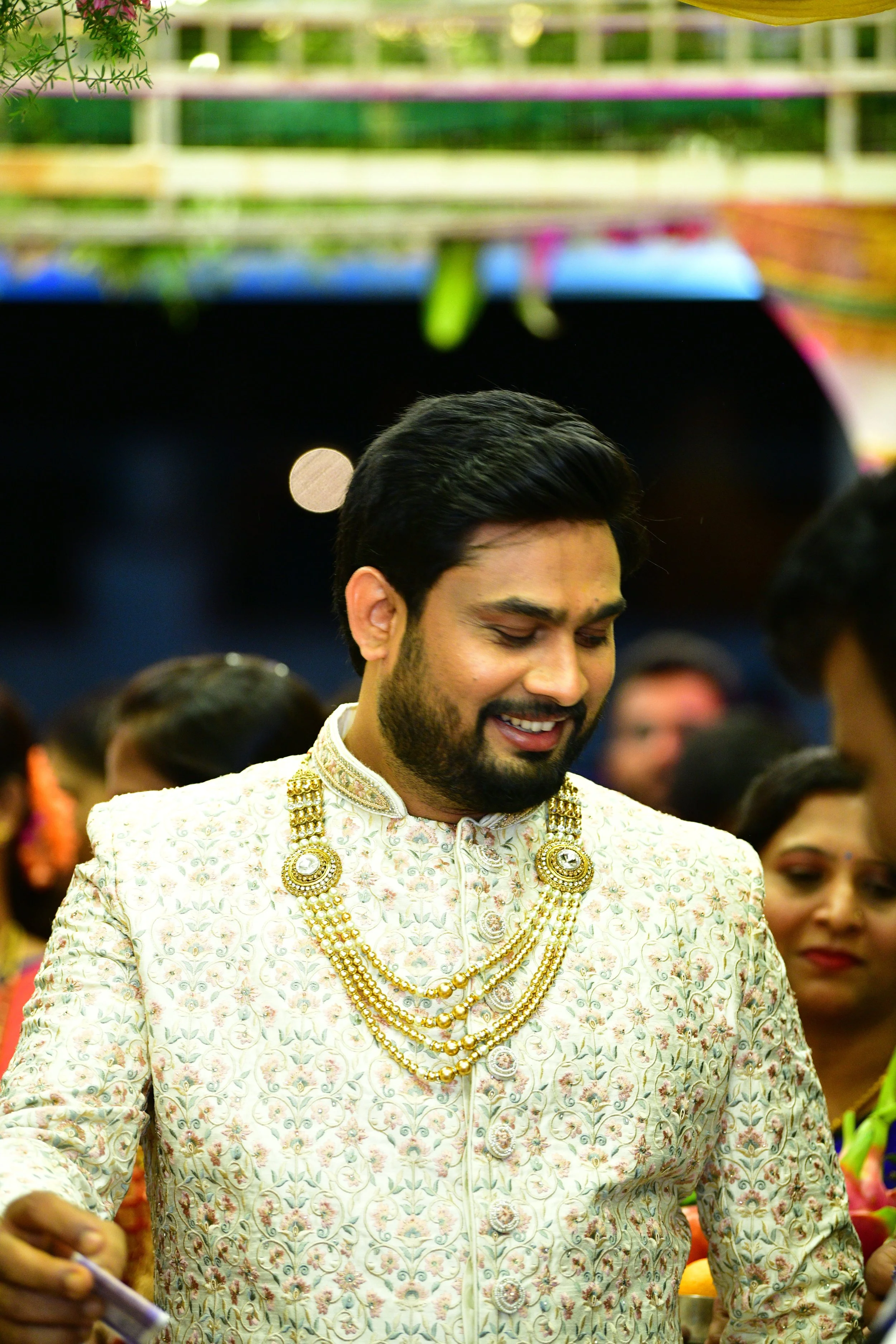 A man dressed in a cream-colored traditional Indian outfit with intricate floral patterns, adorned with layered gold necklaces, at a festive occasion surrounded by other people.
