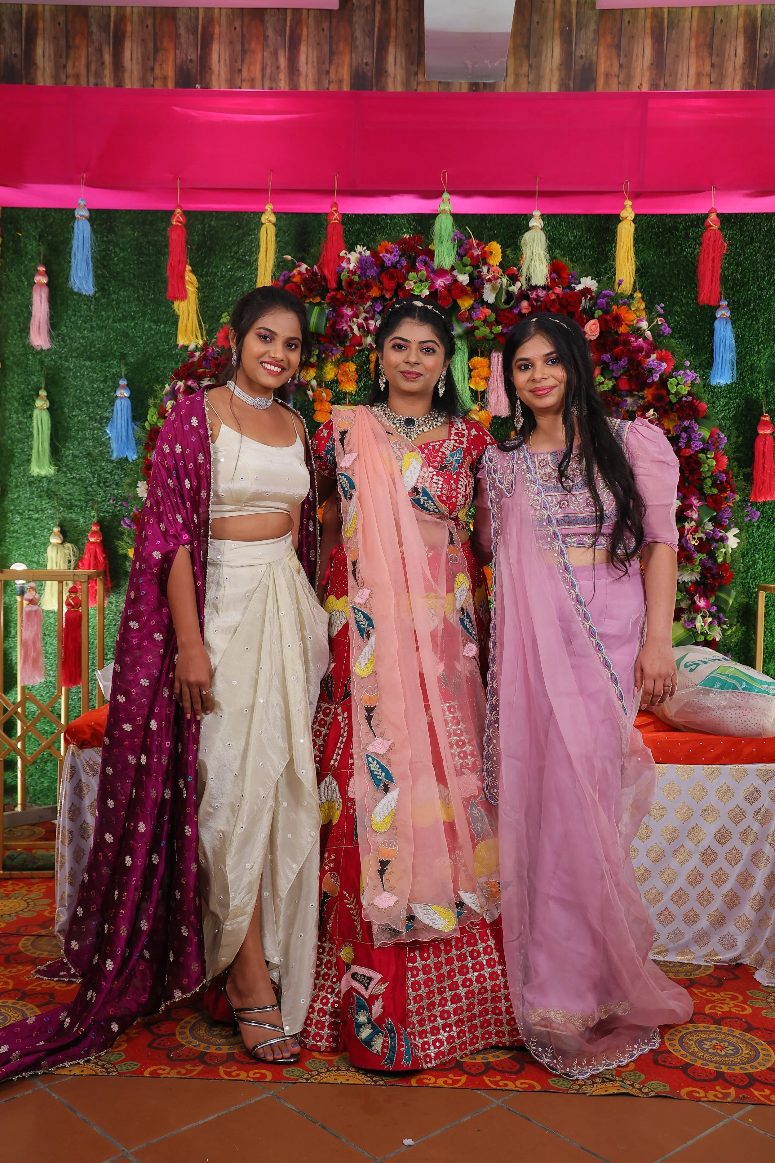 Three women dressed in traditional Indian attire standing in front of a floral backdrop with decorative tassels and a pink fabric awning at a celebration or wedding event.