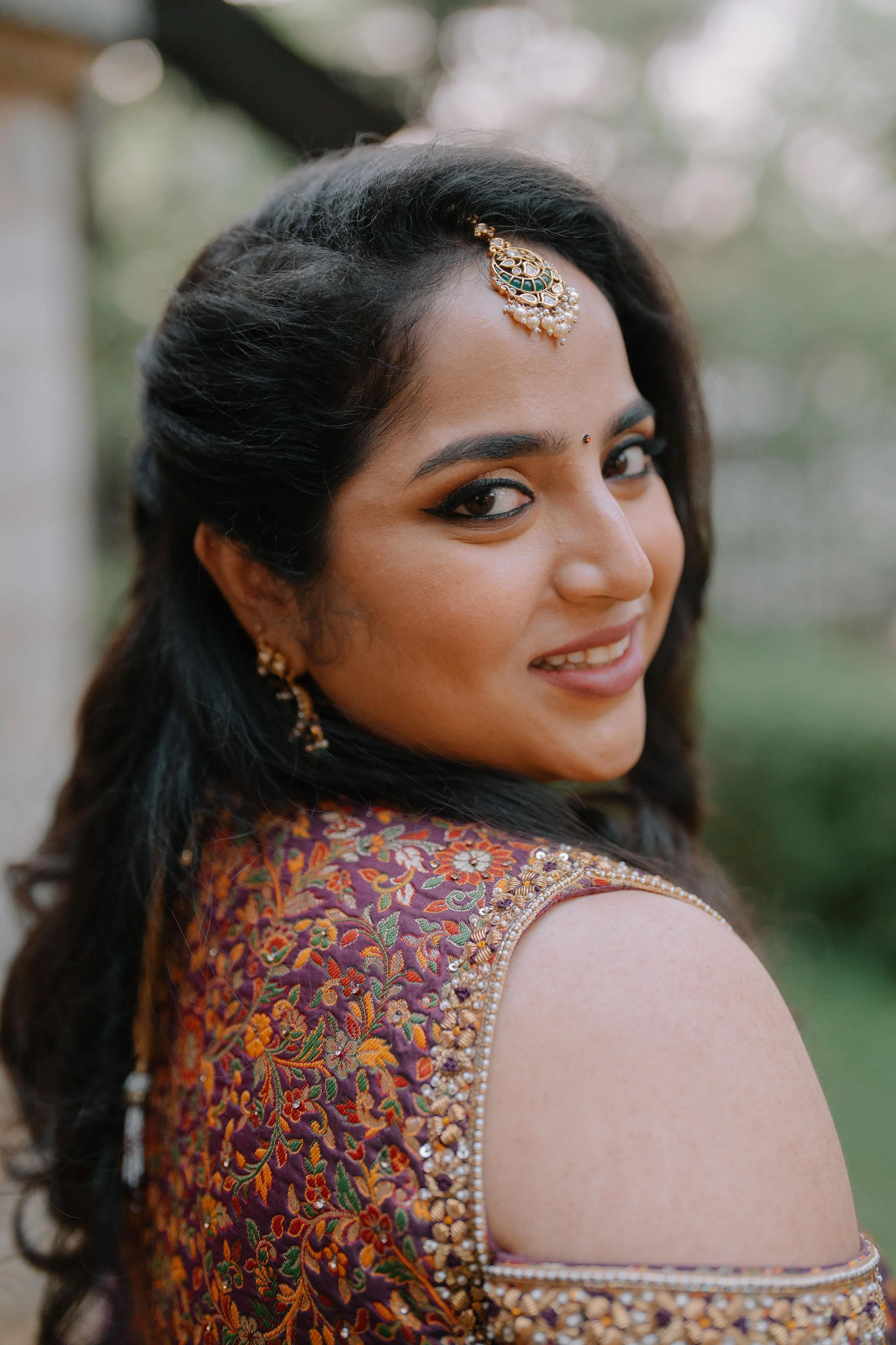 A woman wearing traditional Indian attire, including a richly embroidered blouse with floral patterns and embellishments, with jewelry such as earrings and a maang tikka, smiling outdoors.