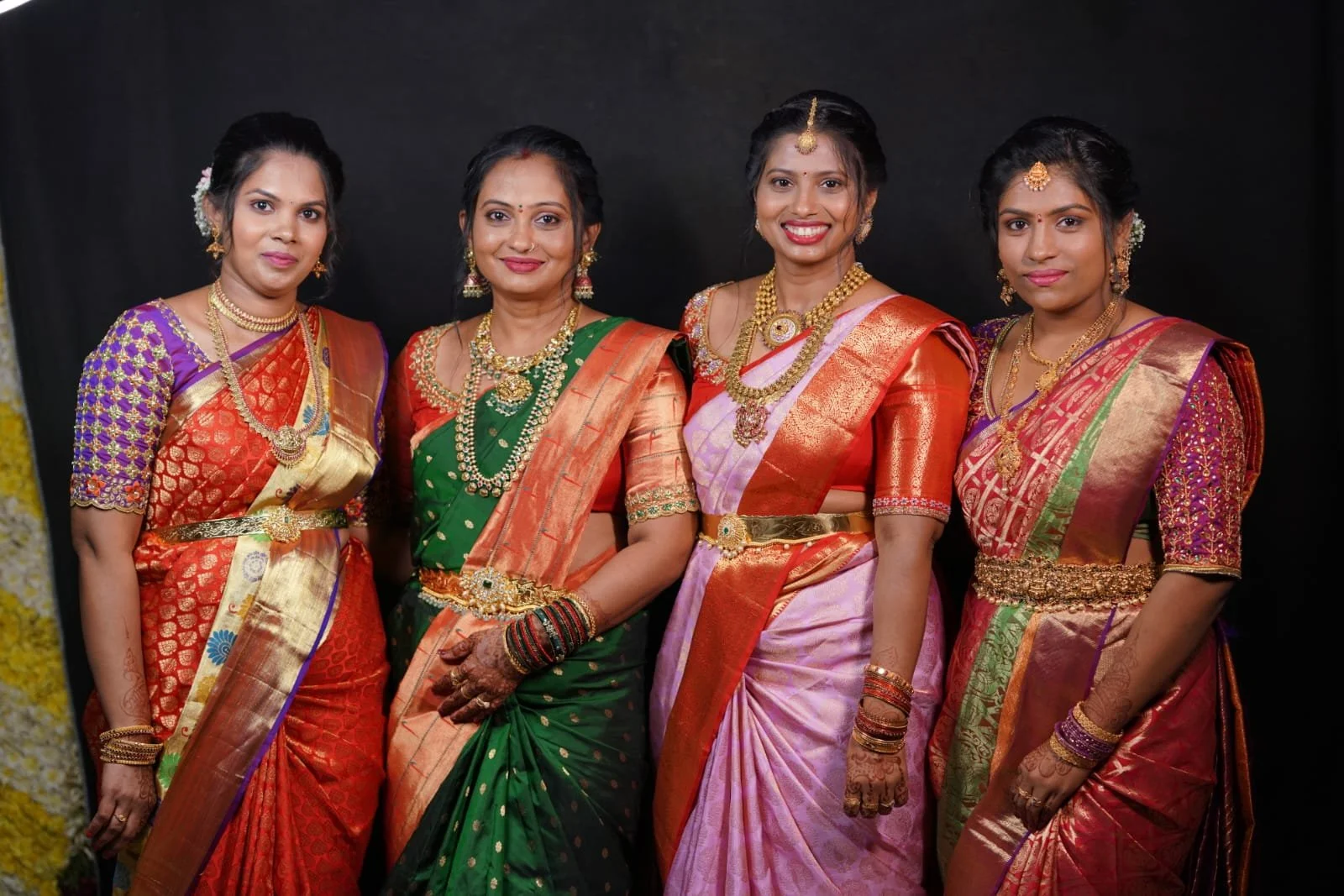 Five women dressed in traditional Indian sarees, wearing jewelry and posing together against a black backdrop.