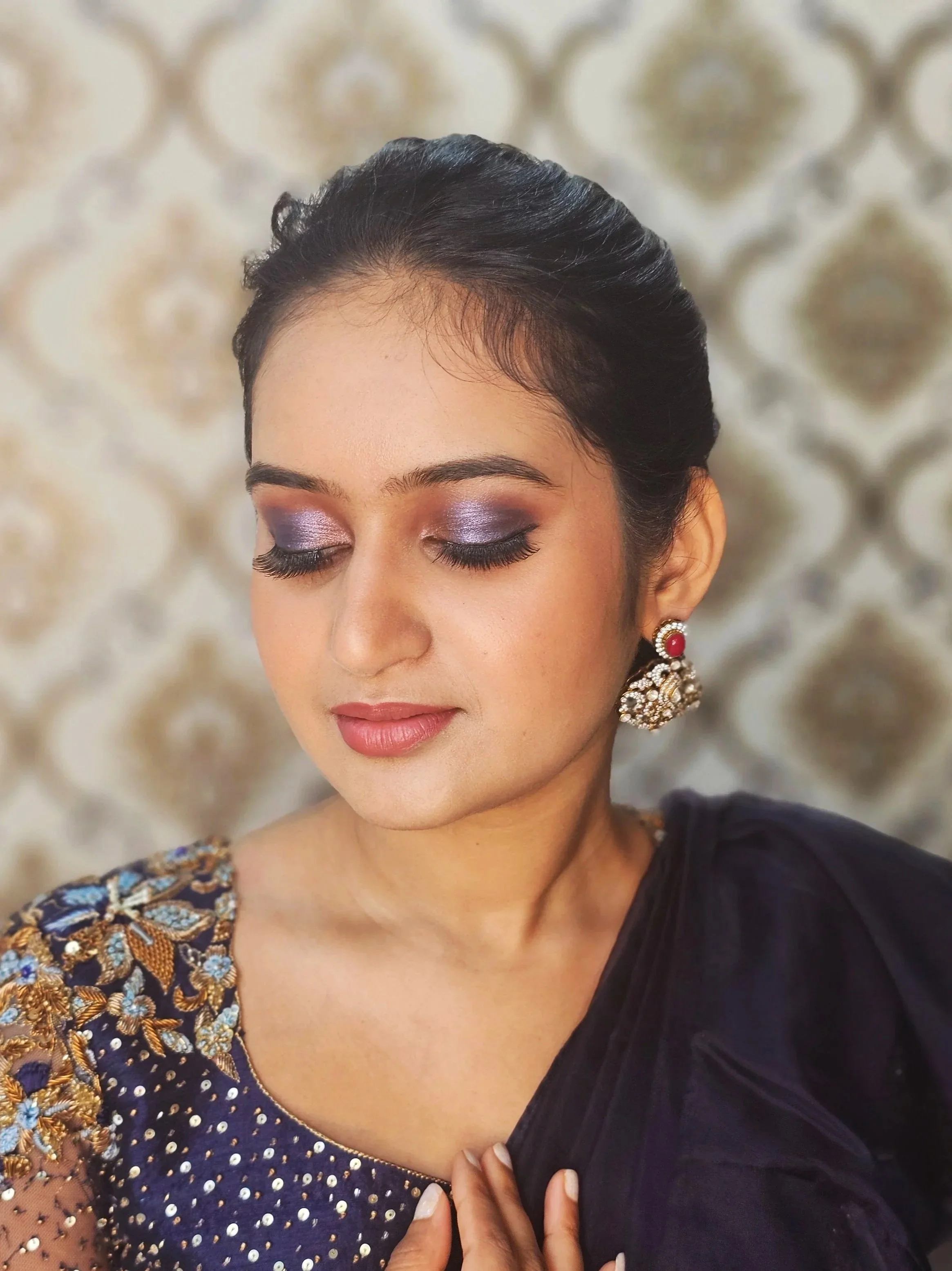 A woman with dark hair styled in an elegant updo, wearing purple eyeshadow, mascara, and pink lipstick, dressed in a navy blue outfit with gold embroidery, and large ornate earrings, standing against a patterned background.