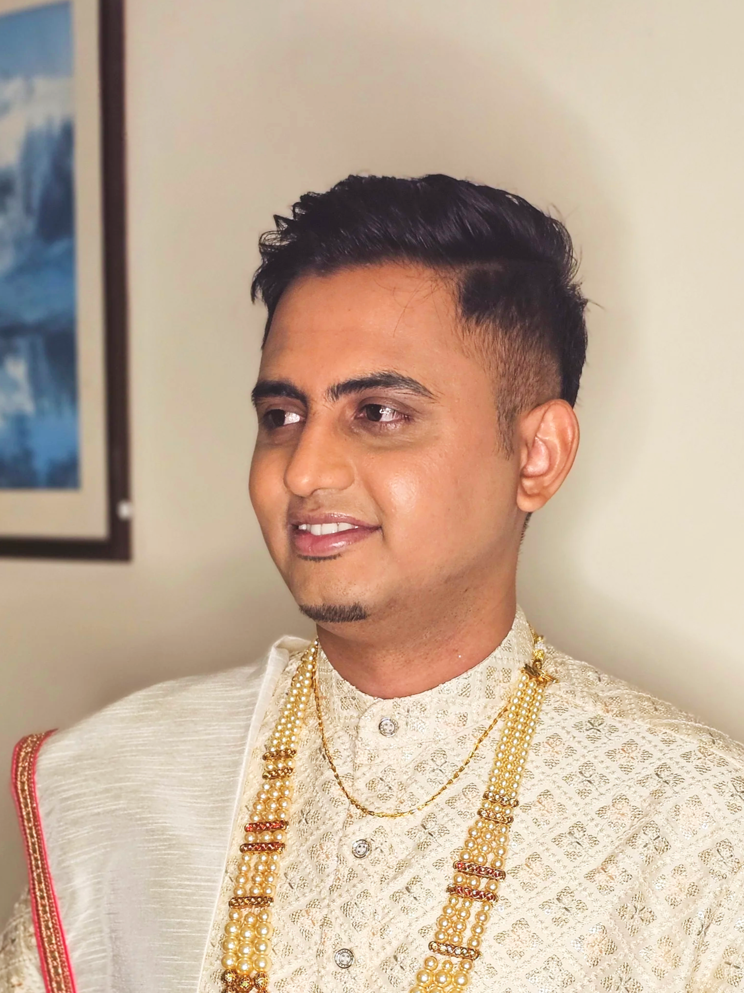A man dressed in traditional Indian attire with ornate jewelry, smiling slightly, standing indoors.