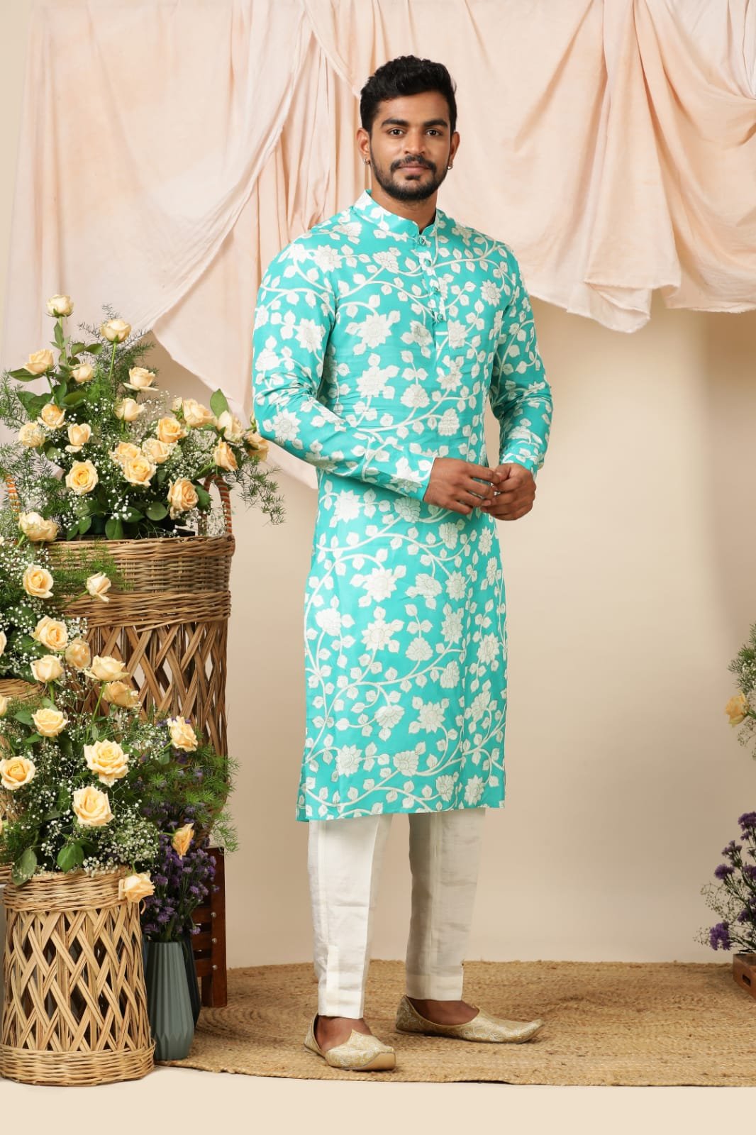 Man wearing a traditional Indian kurta with floral pattern, cream-colored pajama, and cream loafers, standing in front of a beige backdrop with flower arrangements on either side.