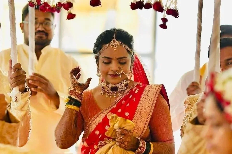 Indian bride wearing red and gold saree, traditional jewelry, and henna on hands during a wedding ceremony.
