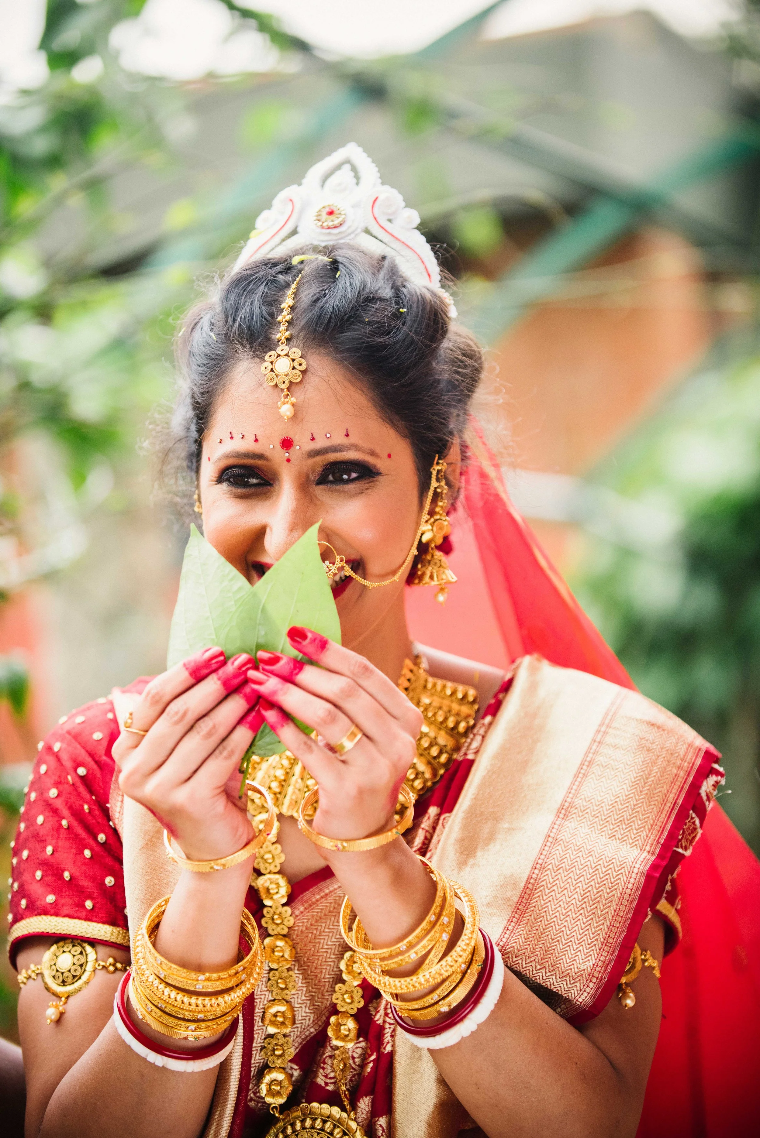 A woman dressed in traditional Indian wedding attire holding green leaves near her nose, smiling with jewelry and red bangles.
