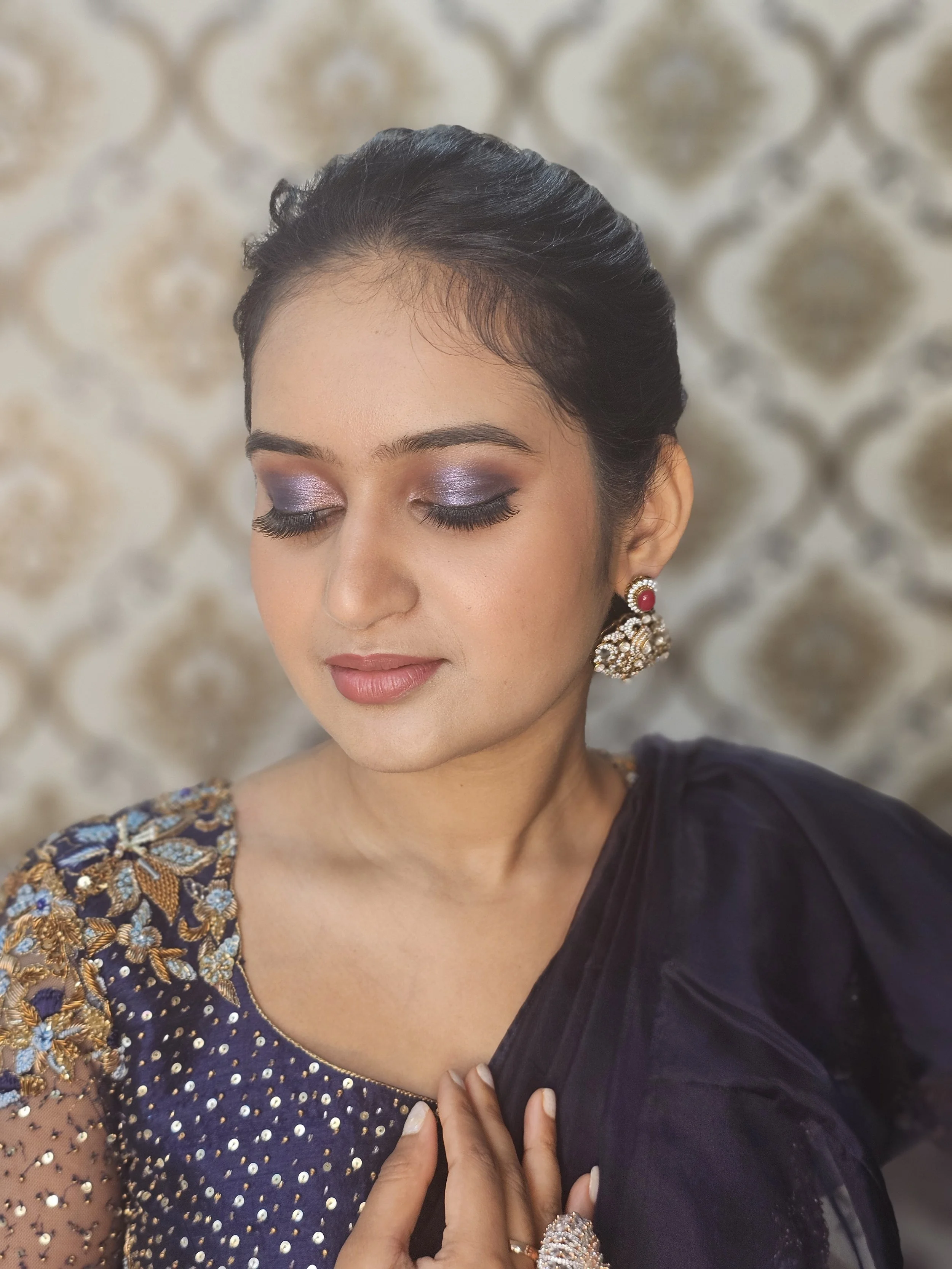 A woman with closed eyes, wearing makeup with purple eyeshadow, gold and blue embroidered attire, large pearl and gold earrings, and a ring, standing in front of a patterned background.