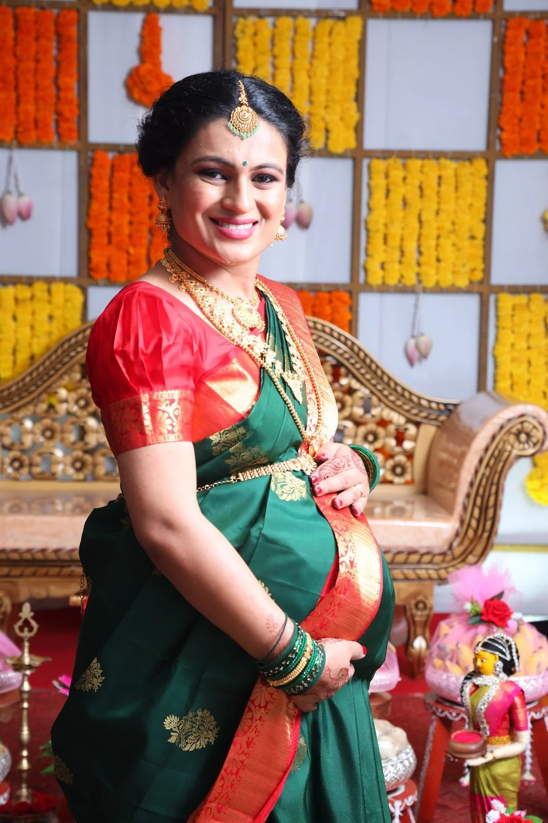Smiling woman in traditional Indian saree and jewelry during a celebration, with flower decorations and a decorative backdrop in the background.