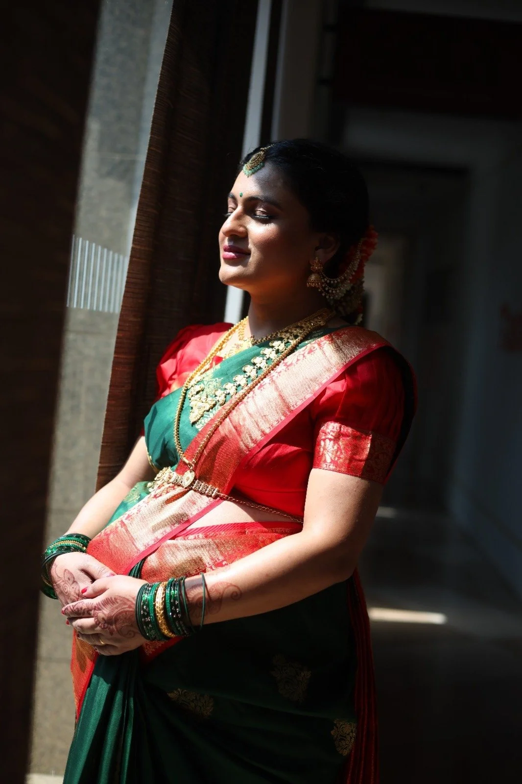 A woman dressed in traditional Indian attire standing near a window with sunlight on her face, wearing a red and green saree with gold jewelry and bangles.