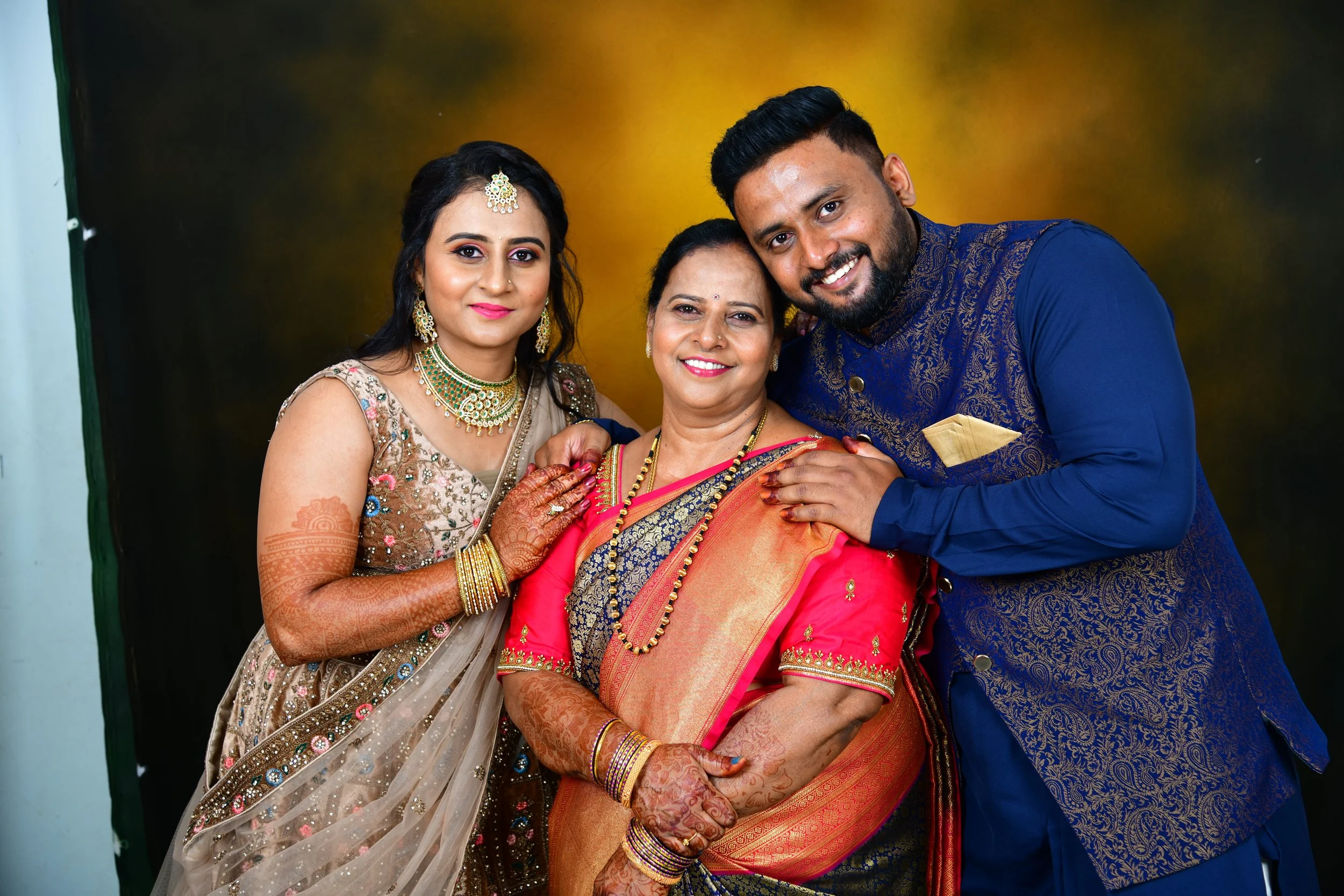 A family of three celebrating with smiling faces, dressed in traditional Indian attire, with a woman in the center wearing a pink saree, a man on the right in a blue kurta with gold designs, and a woman on the left in a beige saree with embroidery.
