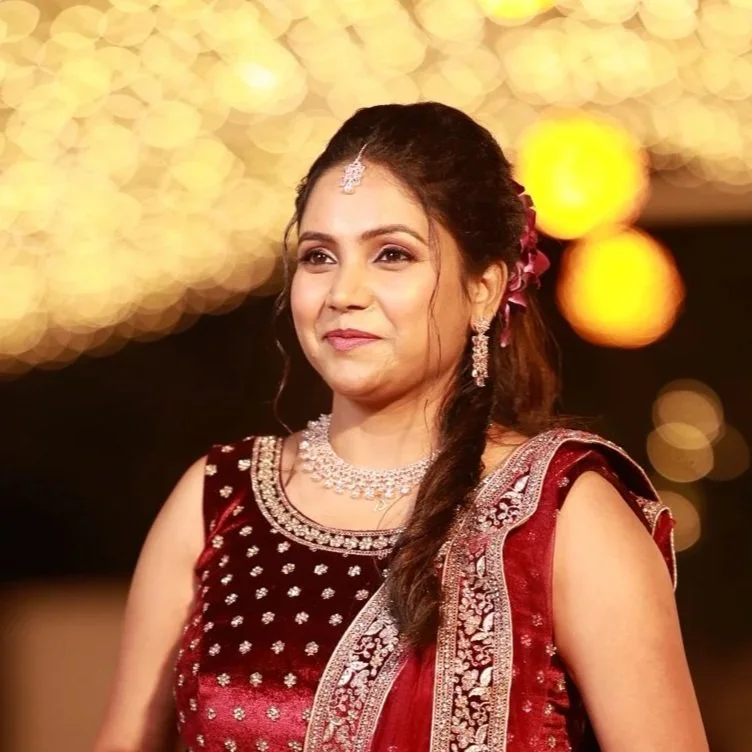 A woman wearing a rich maroon and silver traditional Indian outfit with jewelry, smiling at an event with warm, blurred lights in the background.