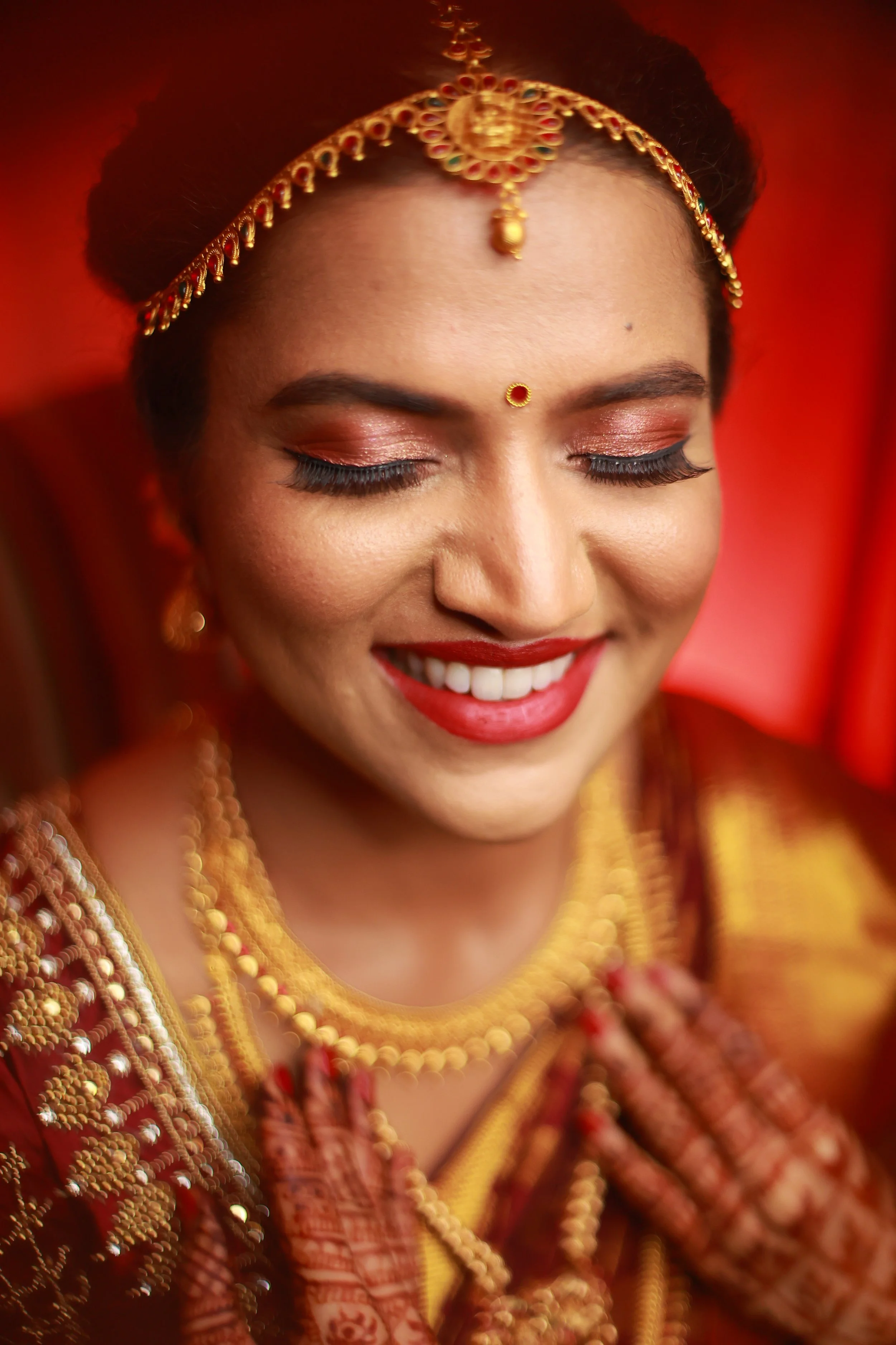 Close-up of a smiling woman dressed in traditional Indian bridal attire, with gold jewelry, red and gold clothing, and henna tattoos on her hands, against a red background.
