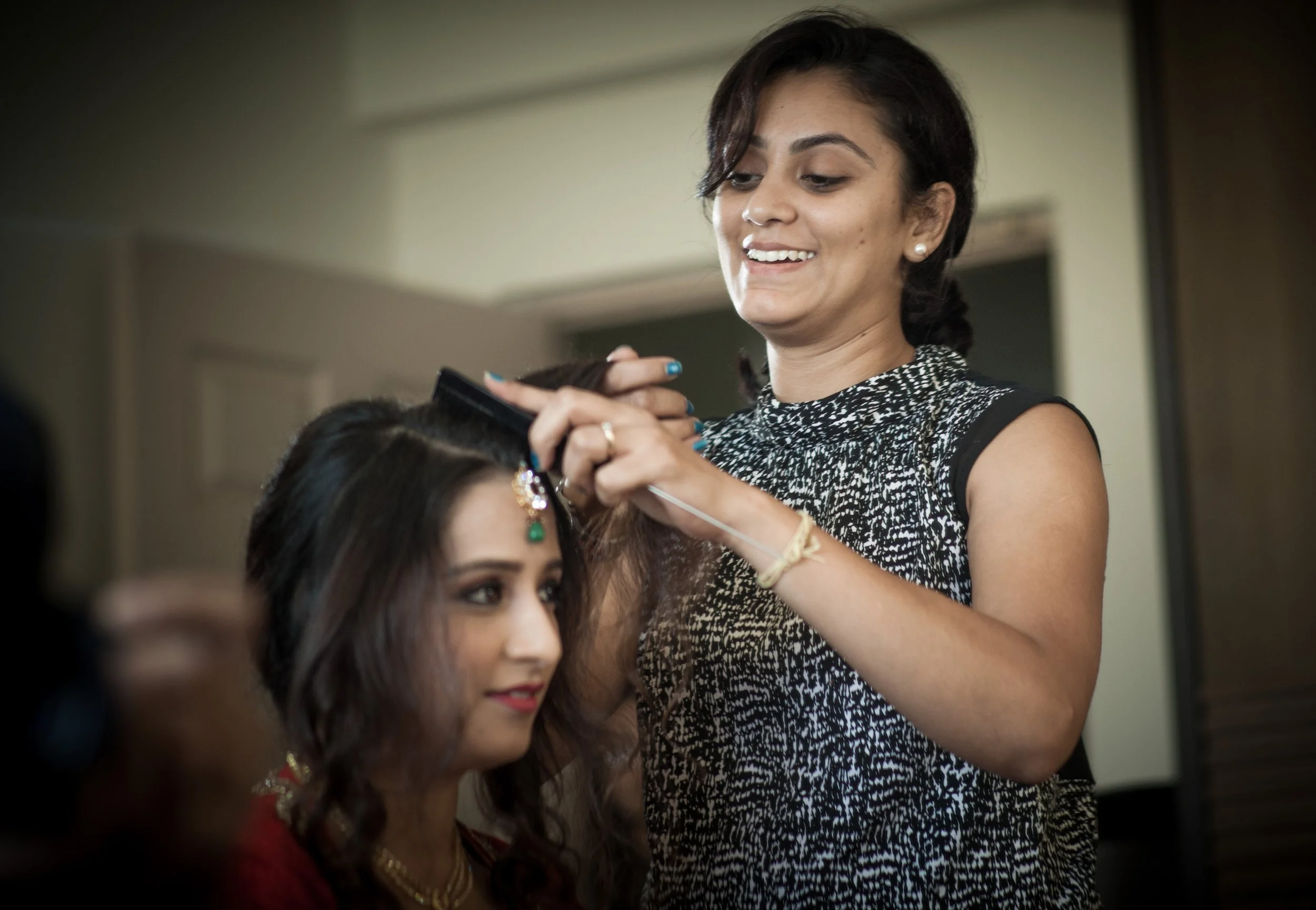 A woman with short dark hair, wearing a sleeveless black and white patterned dress, is styling the hair of another woman with long, dark, wavy hair. The second woman is wearing traditional jewelry, including a maang tikka, earrings, and necklace, and has red lipstick on.