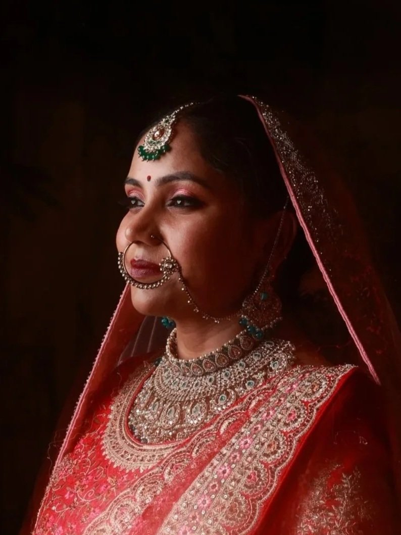 Portrait of a woman dressed in traditional Indian bridal attire with elaborate jewelry, including a maang tikka, nose ring, necklace, and earrings, wearing a red saree with gold embroidery and a veil.