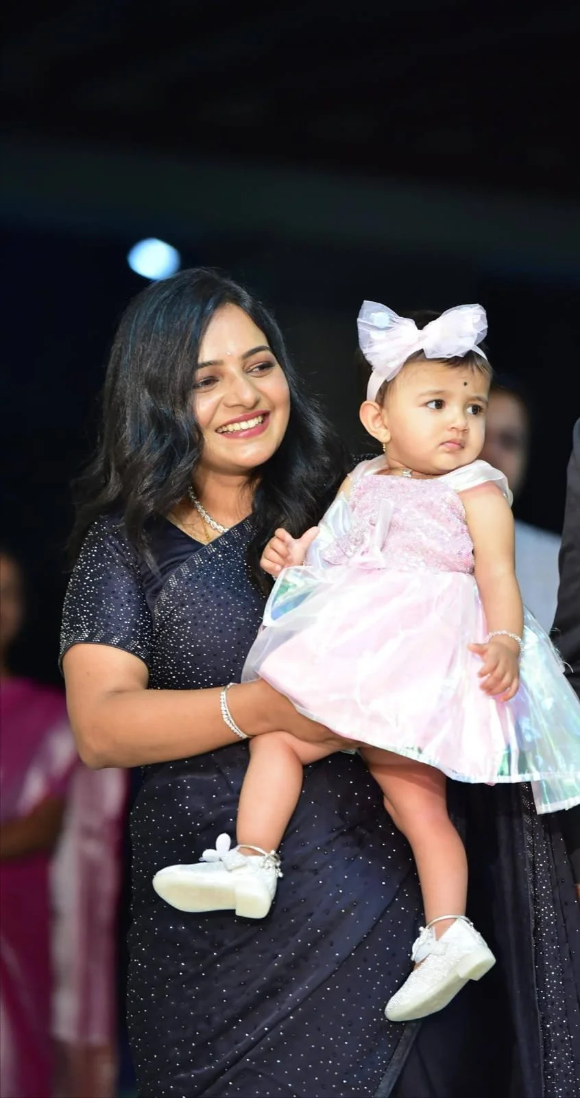 A woman with black hair and a black saree with silver details holding a young girl dressed in a pink and white dress with a large white bow on her head at a celebration or event.