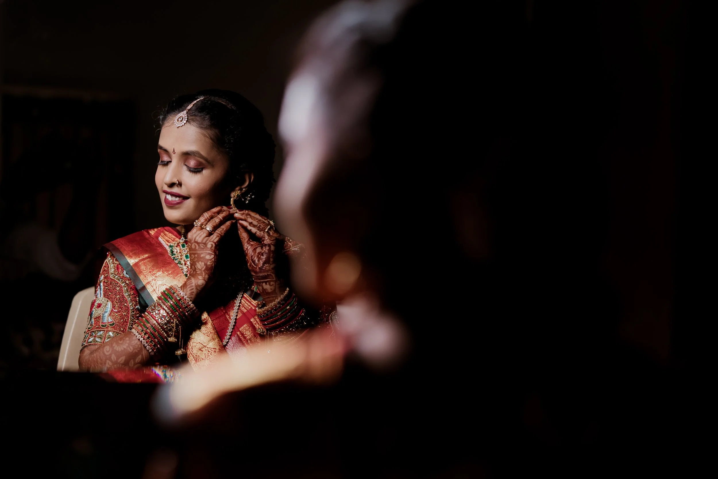 Woman dressed in traditional Indian bridal attire with jewelry, admiring herself and smiling, with out-of-focus person in the foreground