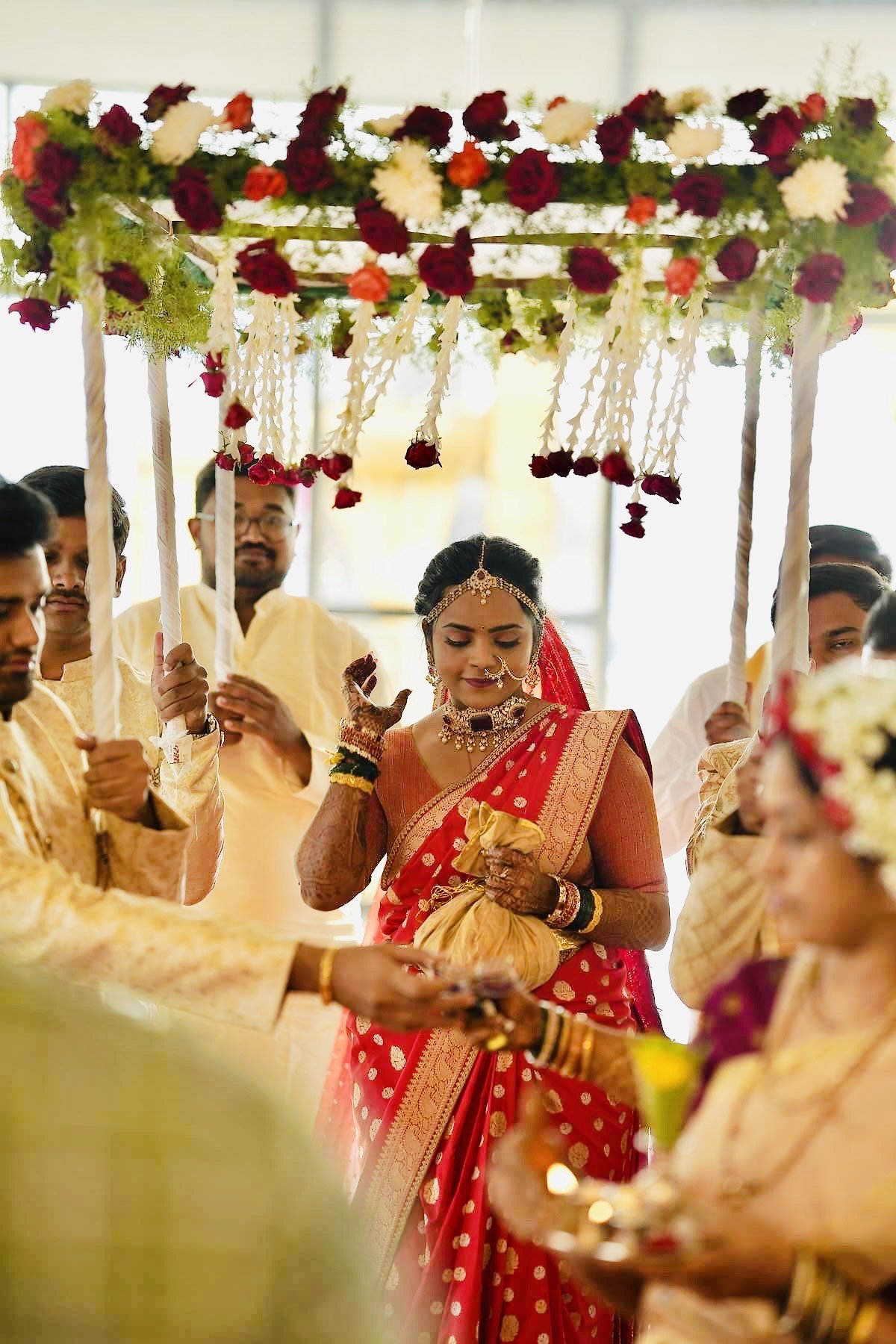 Indian wedding ceremony with a bride in a red and gold saree, surrounded by guests in traditional attire, under a floral canopy decorated with roses and hanging flowers.