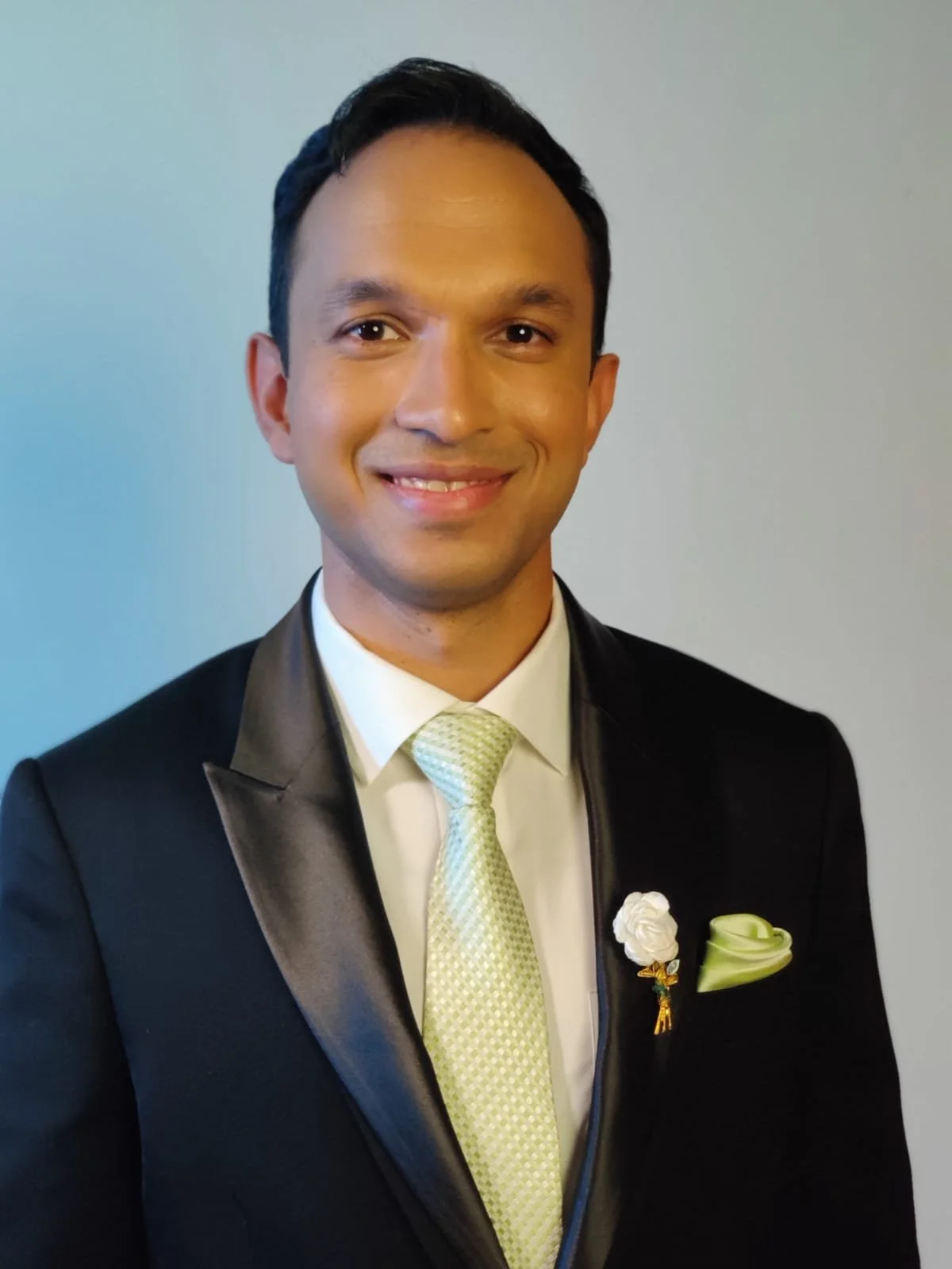 A man in formal attire wearing a black tuxedo with a satin lapel, a white shirt, a light green patterned tie, and a matching pocket square. He has short dark hair and is smiling at the camera against a plain background.