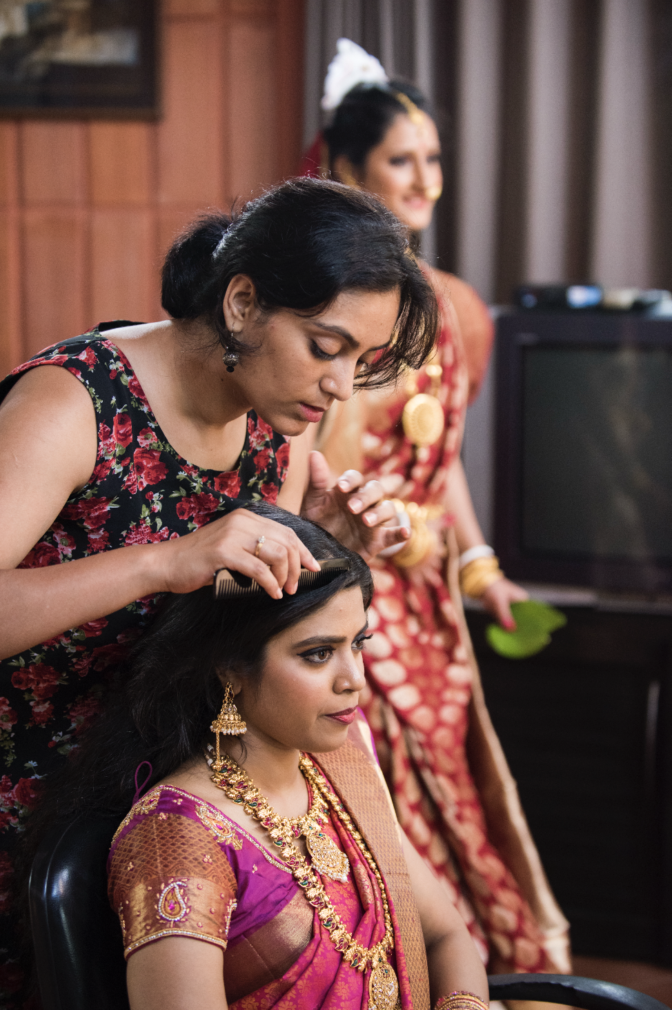 A woman in traditional Indian jewelry and attire sits while another woman styles her hair during a cultural event.