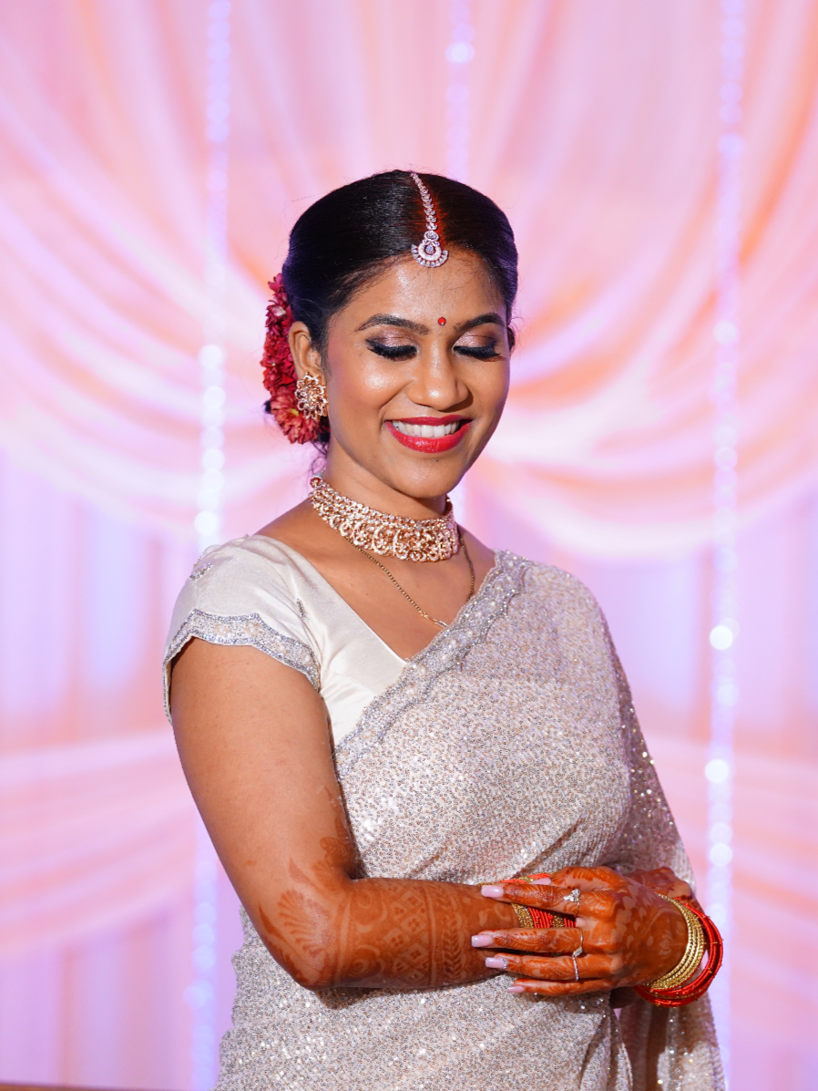 A woman dressed in traditional Indian attire at a celebration, smiling with henna designs on her hands, wearing jewelry and a saree with a bright, decorated background.