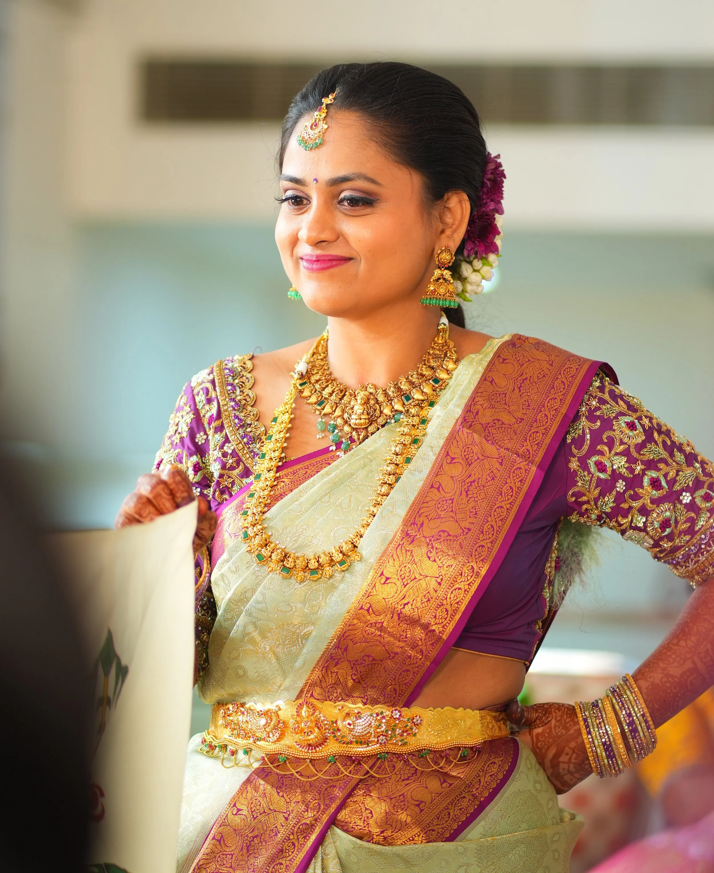 A woman dressed in a traditional Indian saree, adorned with gold jewelry and flowers, smiling softly.