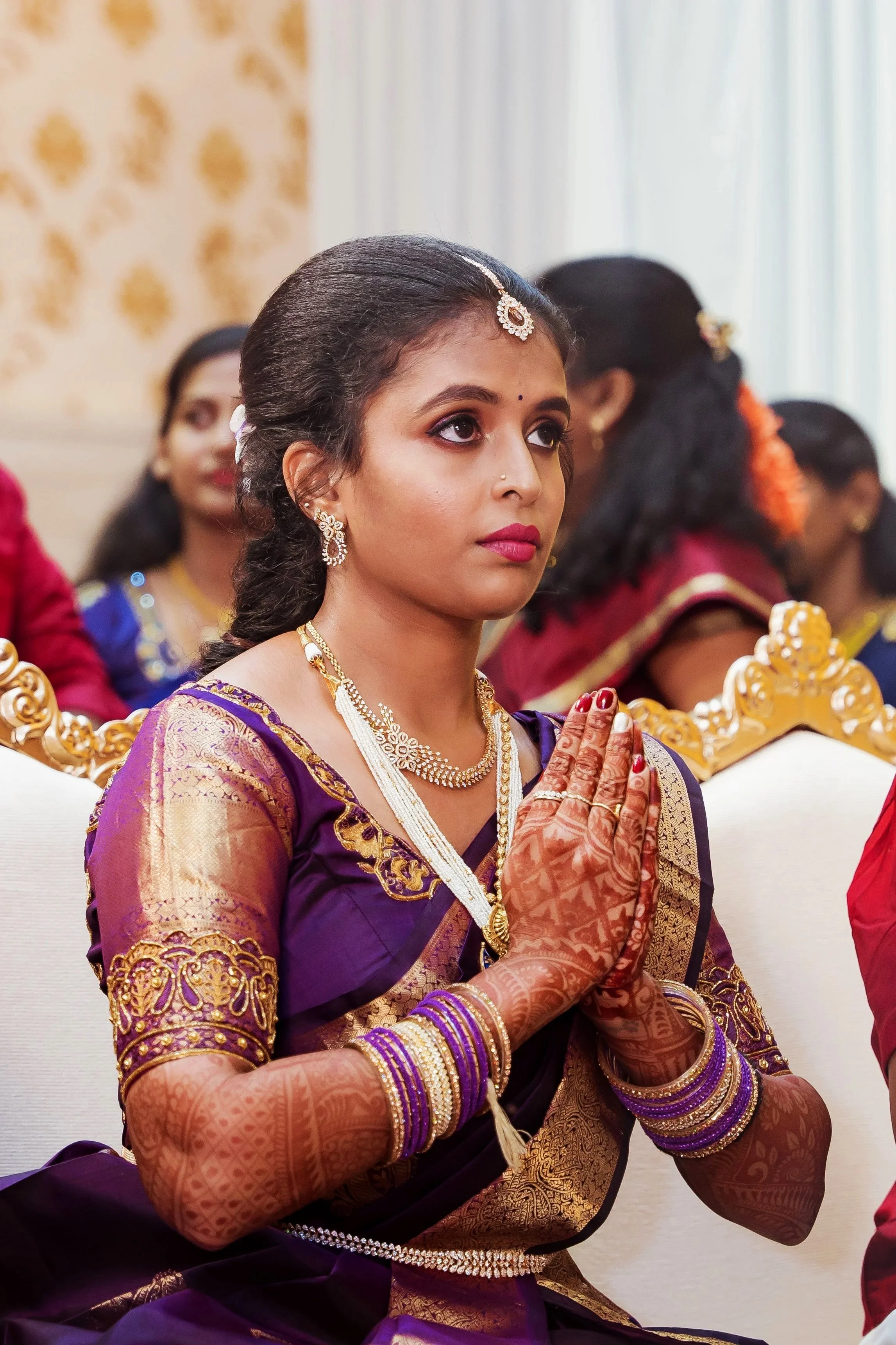 A woman dressed in traditional Indian attire with jewelry and henna on her hands, performing a prayer gesture at a ceremony.