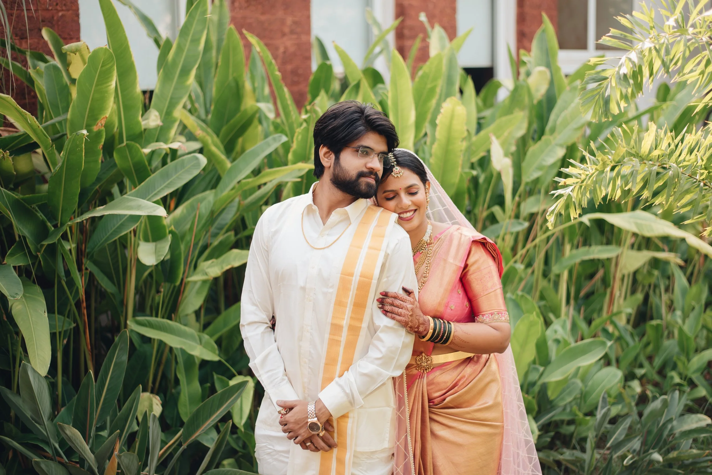 A couple dressed in traditional Indian attire standing closely together among lush green plants, smiling and enjoying each other's company.