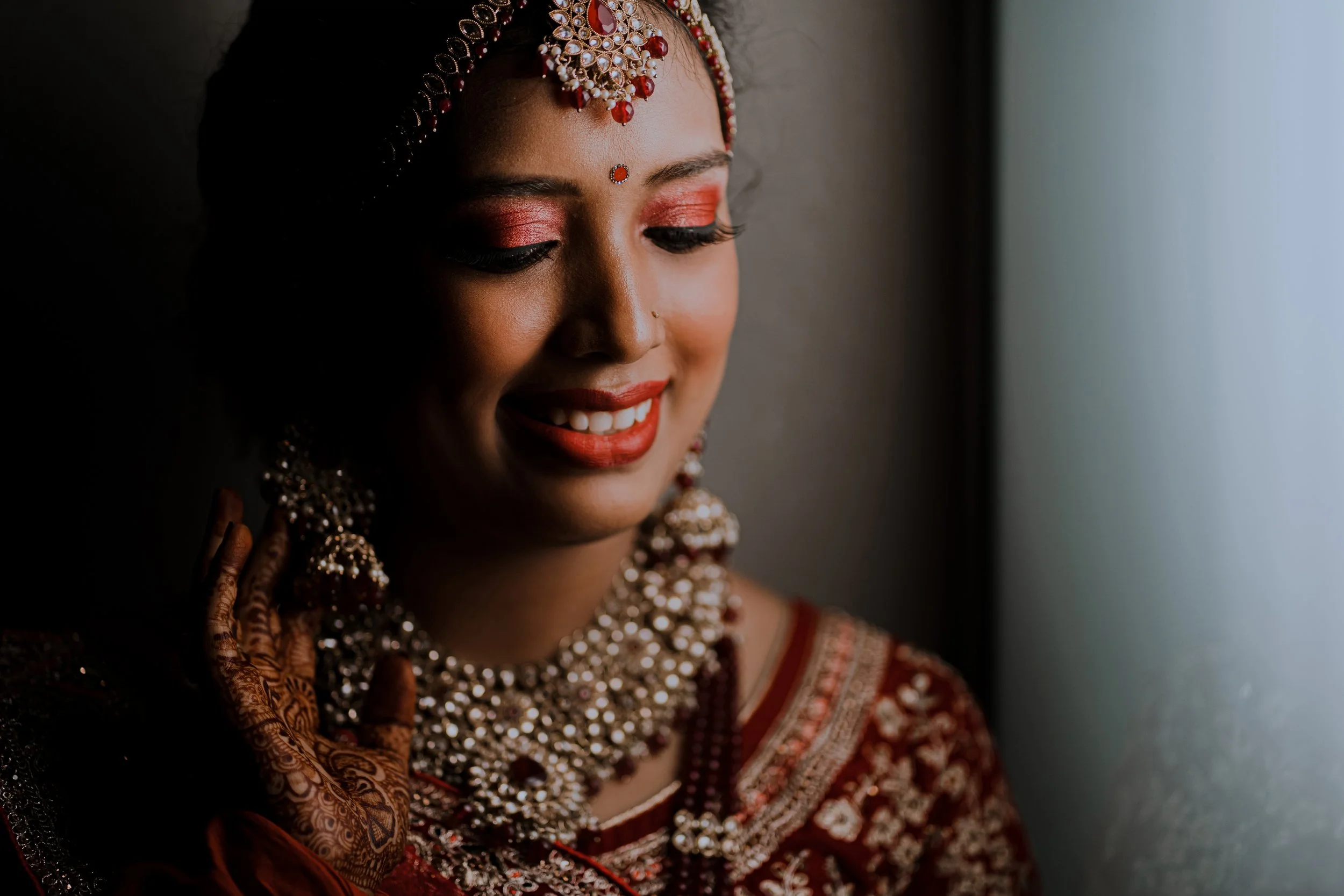 Close-up of a smiling Indian bride with traditional jewelry, henna on her hand, and red makeup, standing near a window.