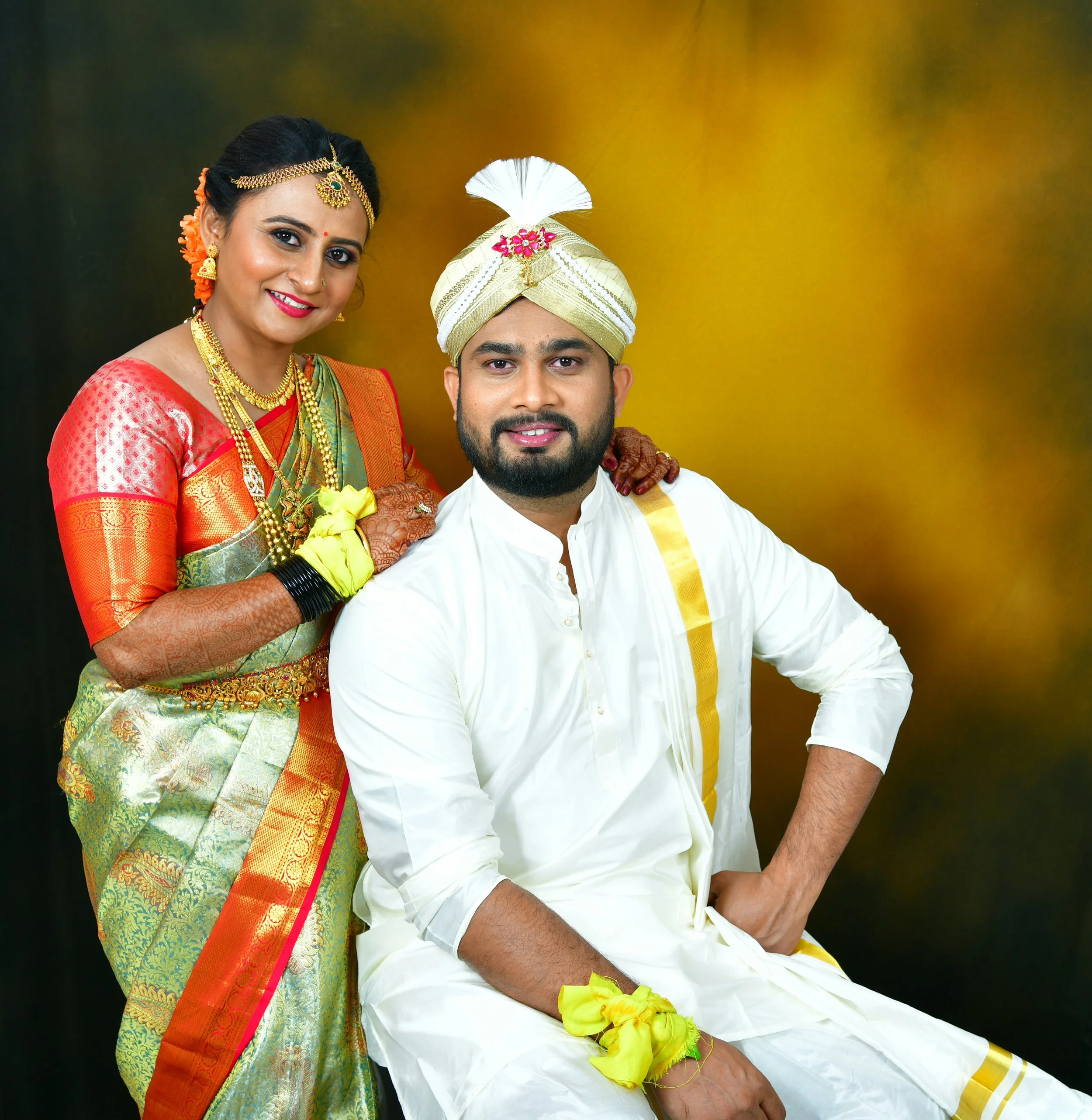 A couple dressed in traditional Indian wedding attire posed against a gold and black background. The woman wears a colorful saree, gold jewelry, and flowers in her hair. The man wears a white kurta with a matching turban adorned with a feather and pi