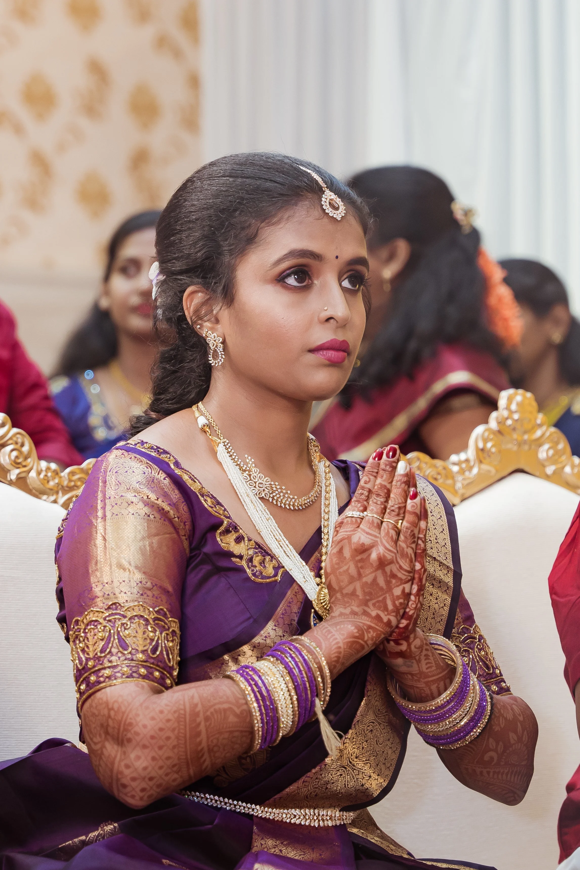 A woman dressed in purple and gold traditional Indian attire, wearing jewelry, and with her hands in a prayer position during a cultural or religious ceremony.