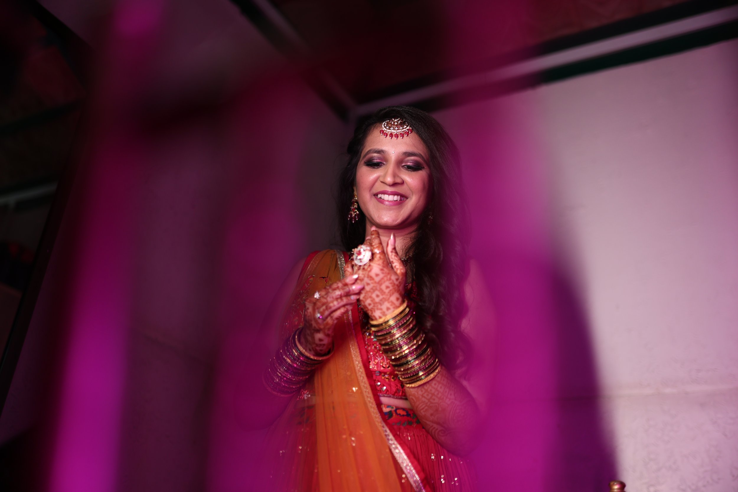 A woman dressed in traditional Indian attire celebrating, smiling, with jewelry and mehndi on her hands, seen through a pink fabric.