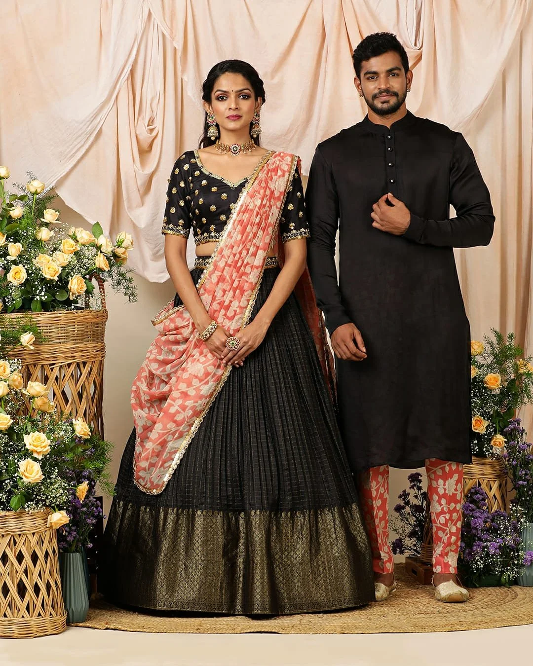 A man and woman dressed in traditional Indian clothing stand in front of a beige curtain backdrop, surrounded by flower arrangements with white, yellow, and purple flowers.