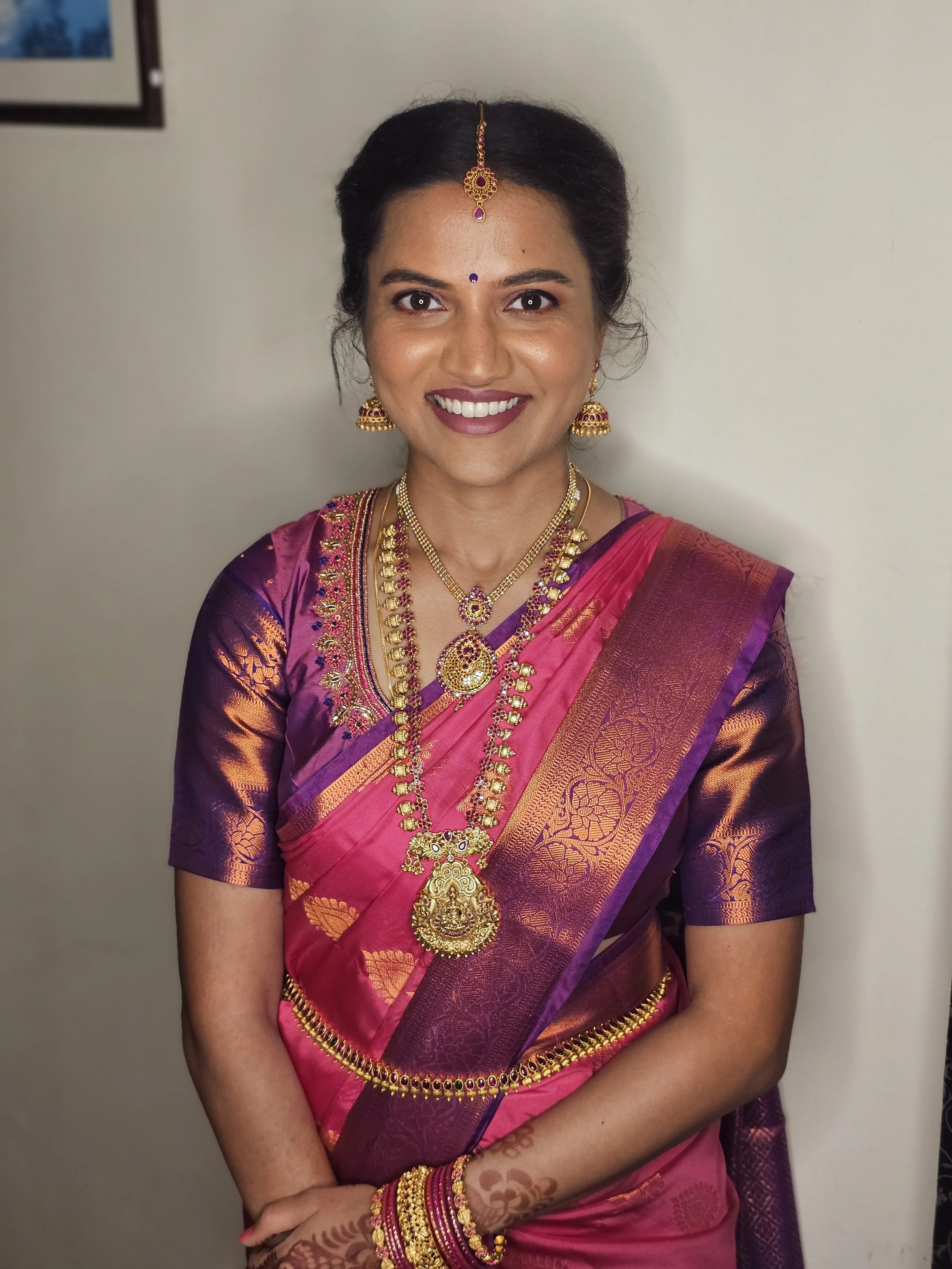 A woman dressed in a pink and purple traditional Indian saree, wearing gold jewelry and smiling.