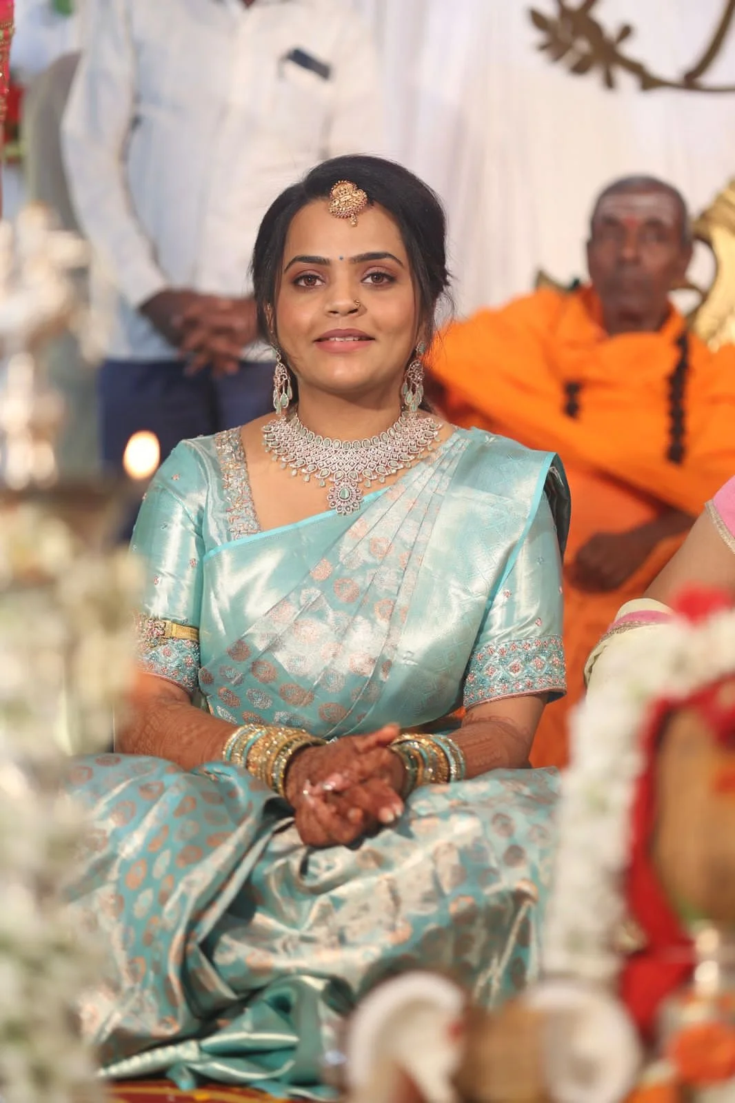 A woman dressed in a traditional light blue saree with silver accents, wearing jewelry including earrings, necklace, bangles, and a headpiece, sitting during a cultural or religious ceremony.