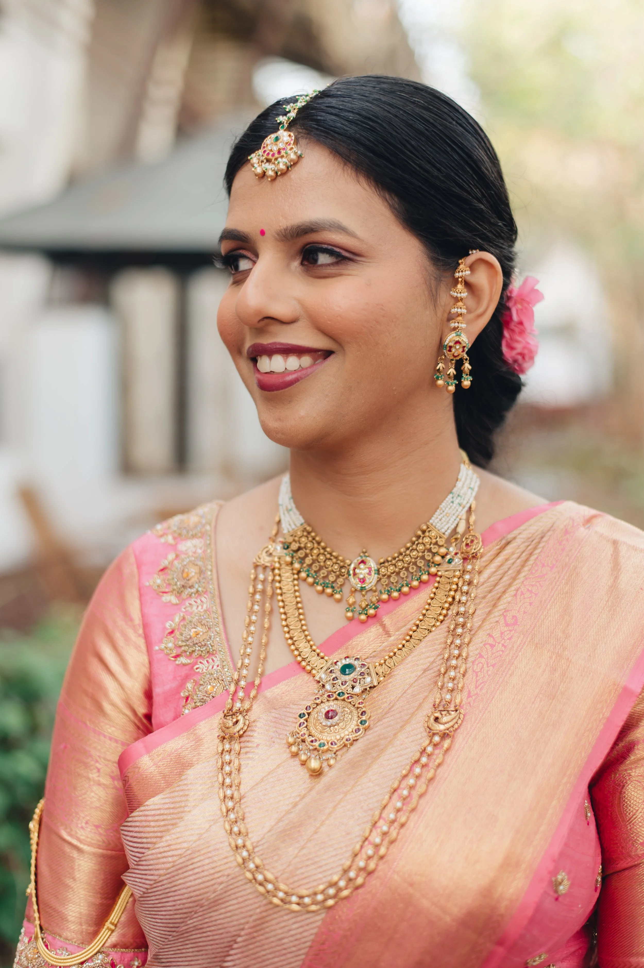 A smiling woman dressed in traditional Indian attire, wearing a pink saree with gold embroidery and adorned with elaborate gold jewelry, including necklaces, earrings, and a headpiece, with her hair styled in a braid decorated with pink flowers.