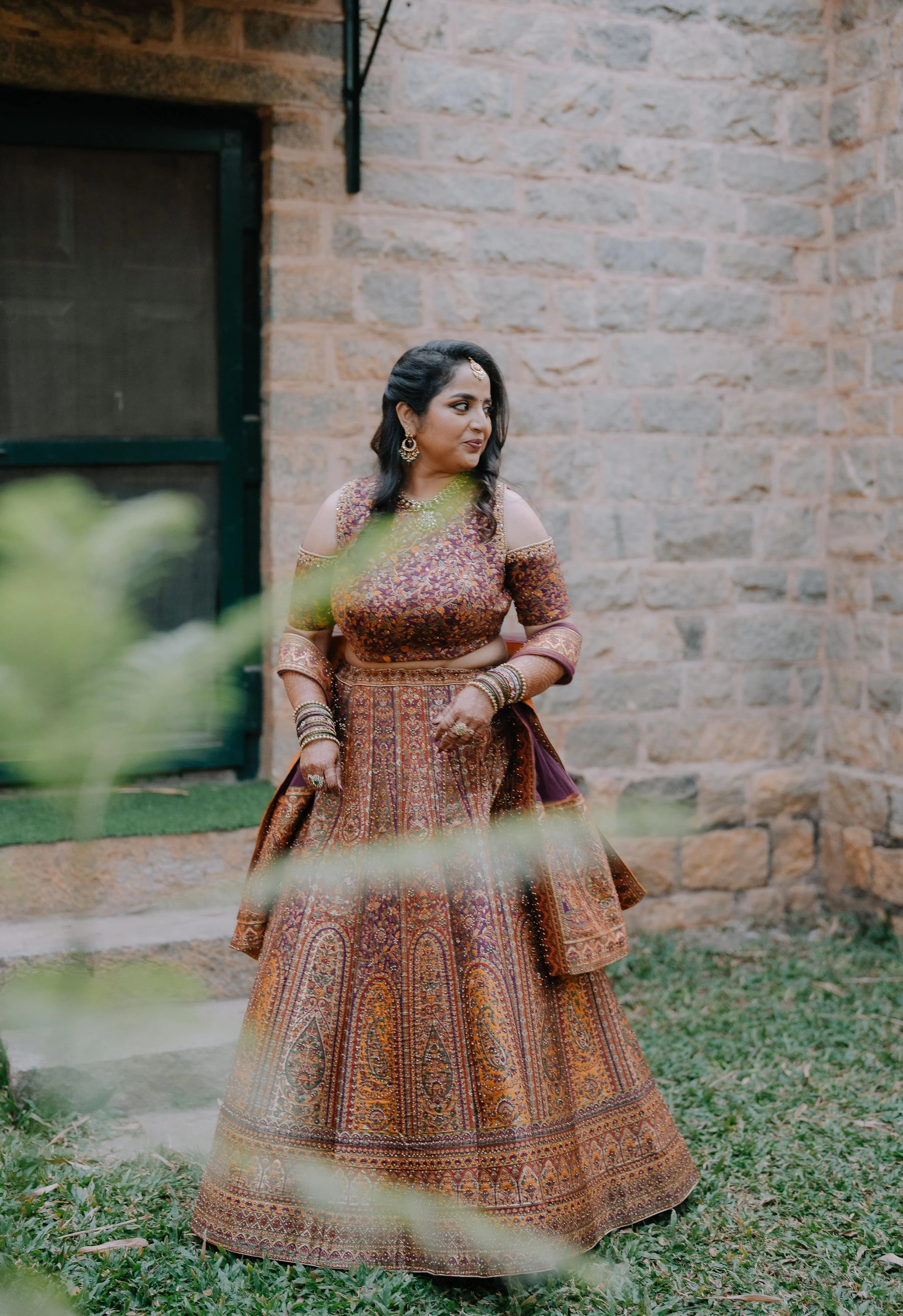 A woman wearing a traditional Indian bridal outfit with intricate embroidery stands outdoors next to a brick building, with greenery and leaves partially obscuring the view.
