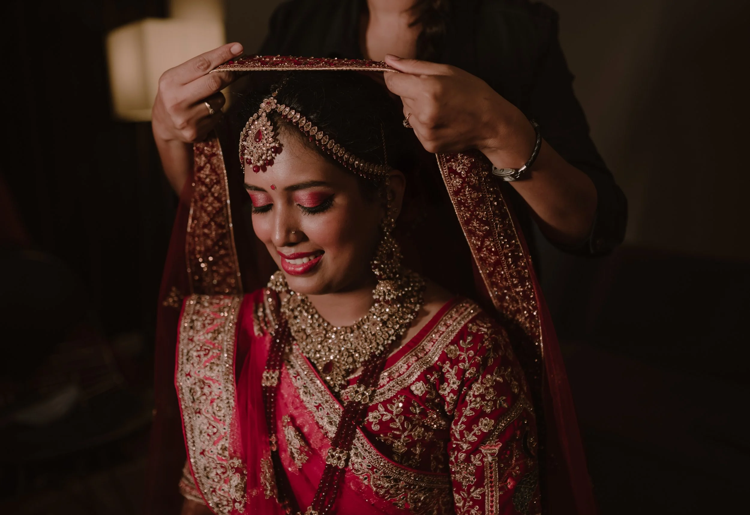 A bride in traditional Indian wedding attire smiling as her veil is being adjusted by another person