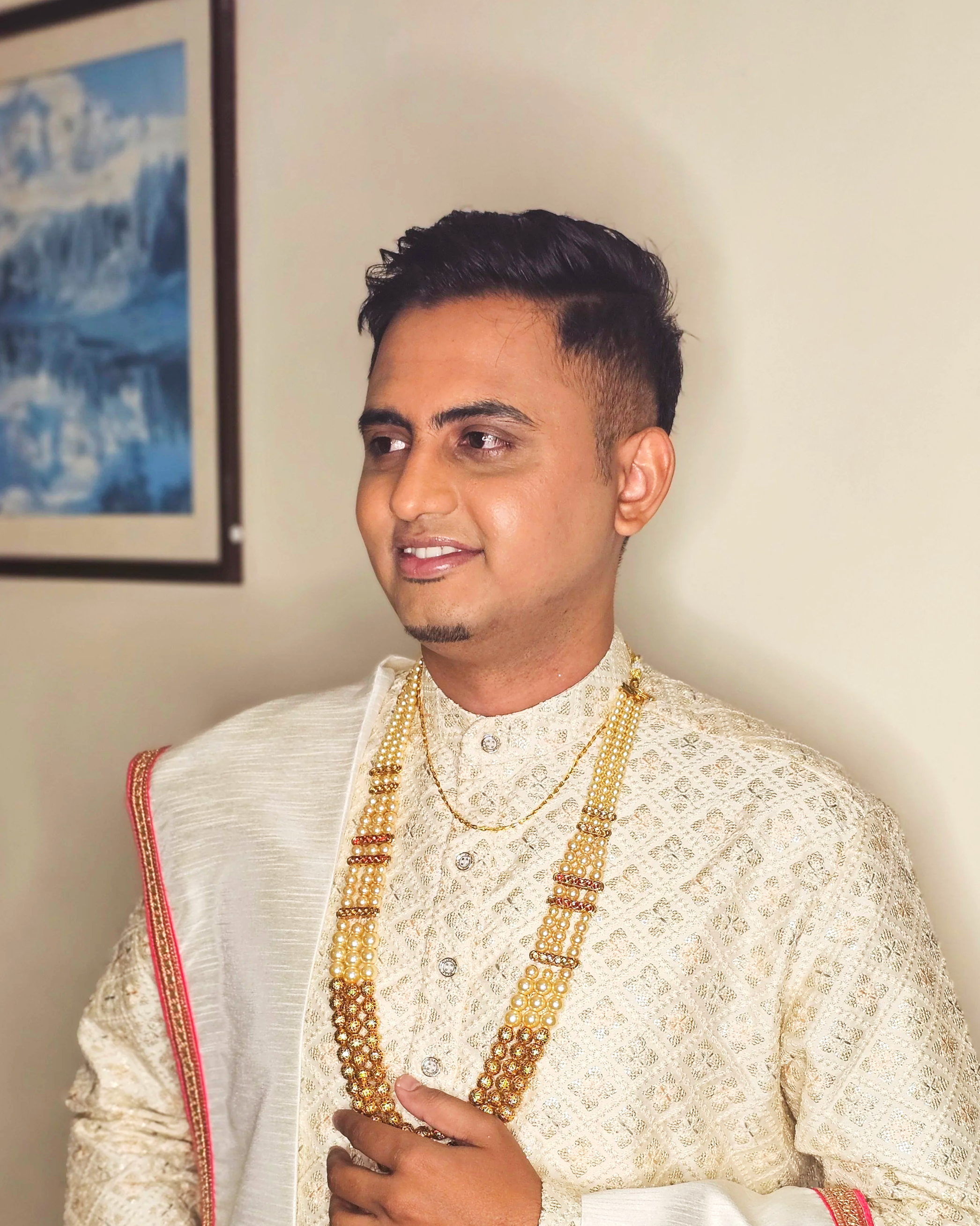 A man wearing traditional Indian wedding attire, including a cream-colored embroidered kurta, gold necklaces, and a cream shawl with red and gold borders, standing indoors near a wall with a framed picture of a waterfall.