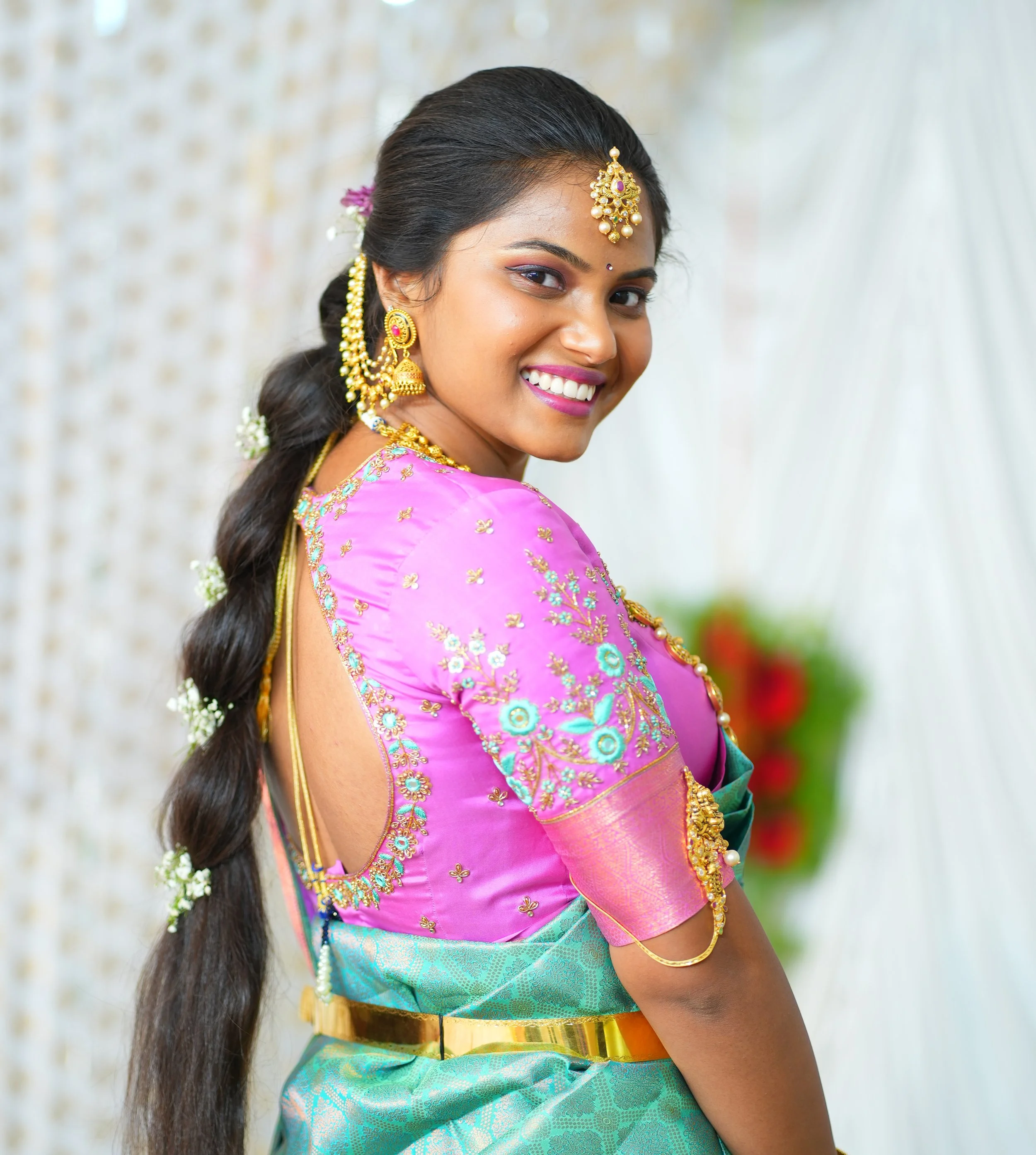 A woman in traditional Indian attire, smiling and looking over her shoulder, wearing a pink and turquoise saree with gold jewelry and flowers in her braided hair.