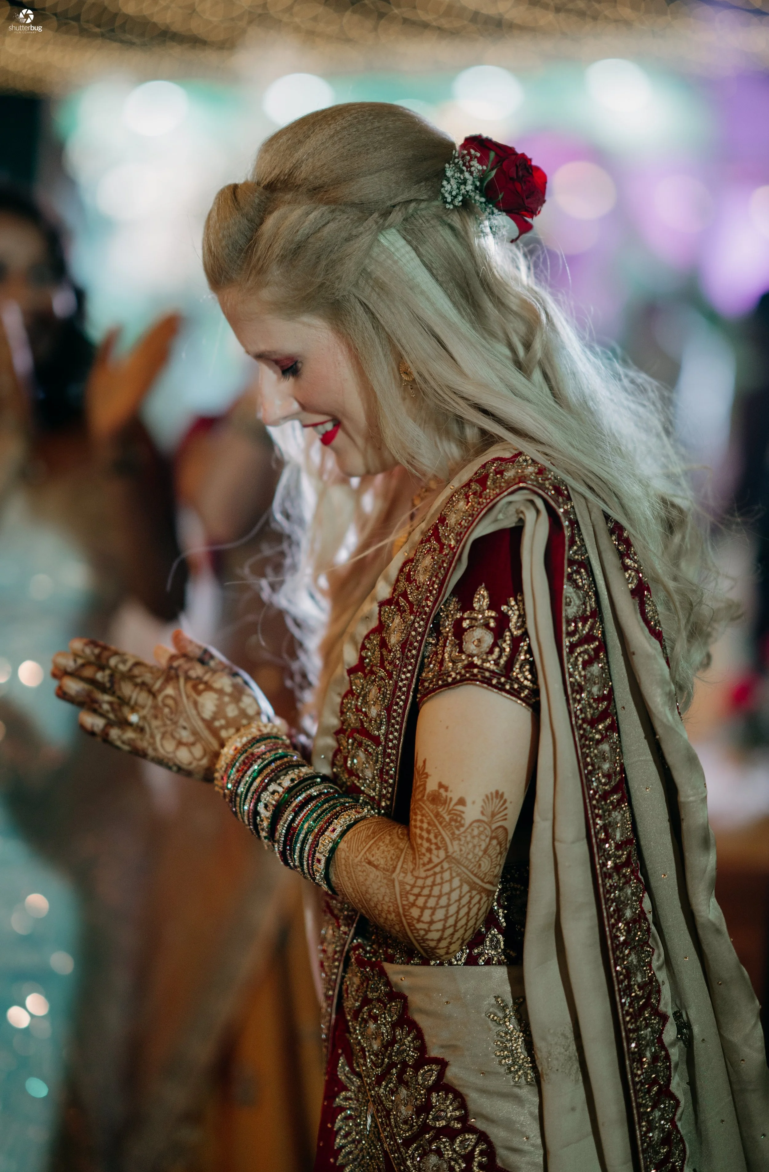 A woman dressed in traditional Indian wedding attire, smiling and praying with her hands folded, wearing intricate henna designs and colorful bangles.