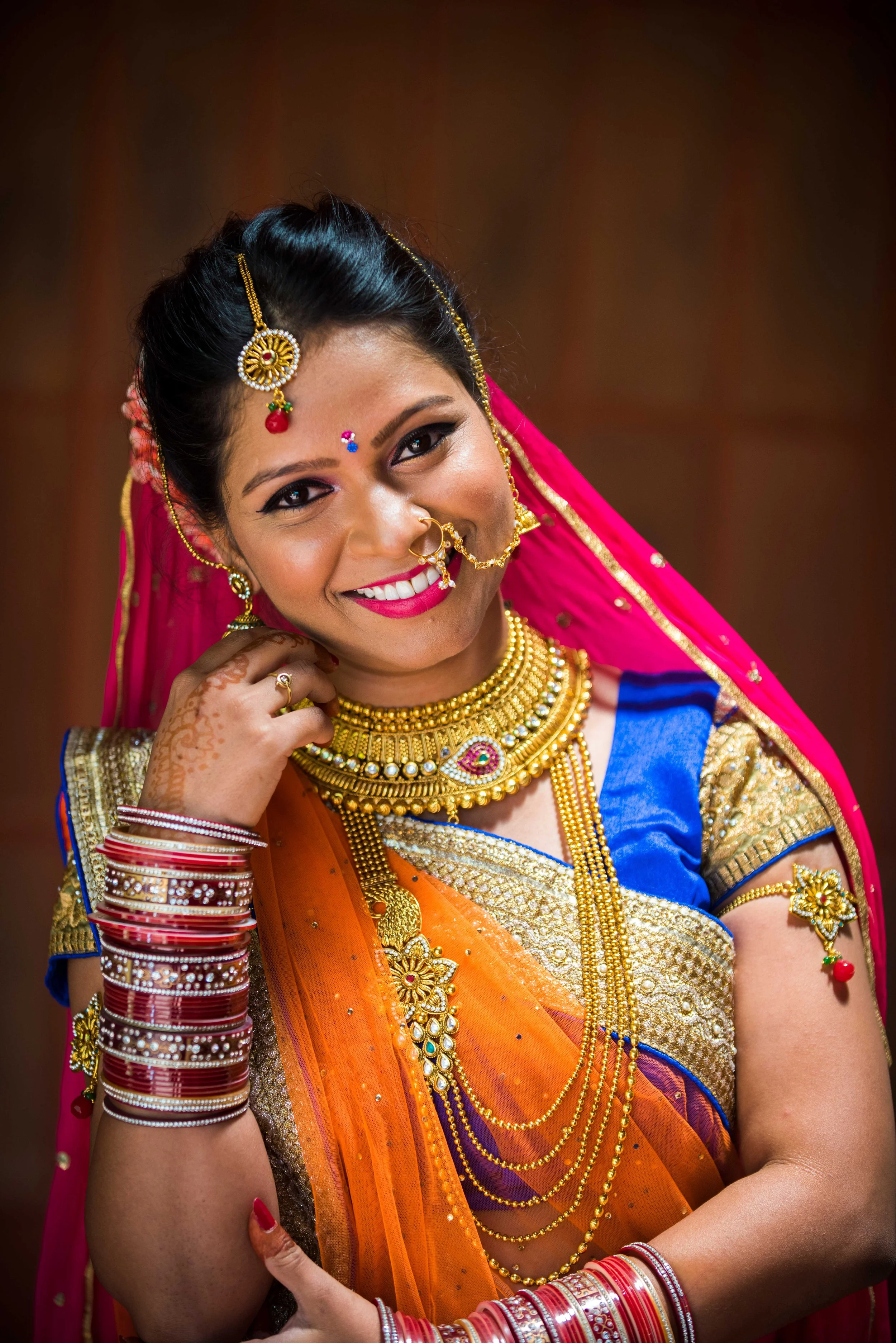 Smiling woman dressed in traditional Indian saree with gold jewelry and red bangles.