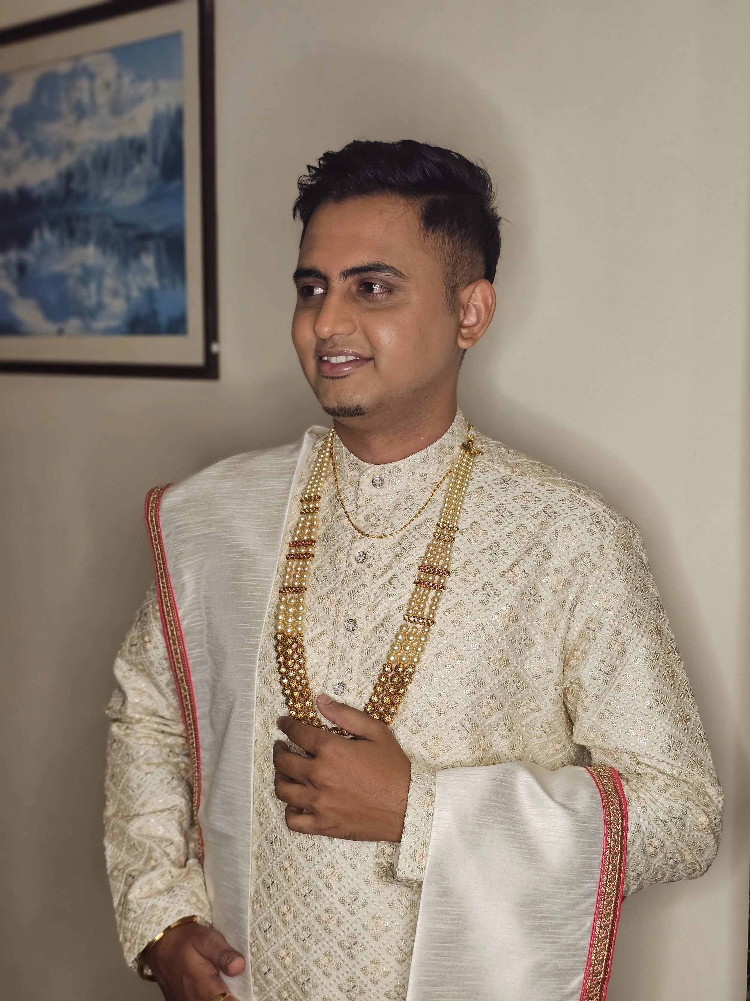 A man in traditional Indian wedding attire, including a cream-colored sherwani with gold embroidery, gold necklaces, and a cream dupatta with red and gold border, standing against a beige wall with a framed picture of mountains and water.